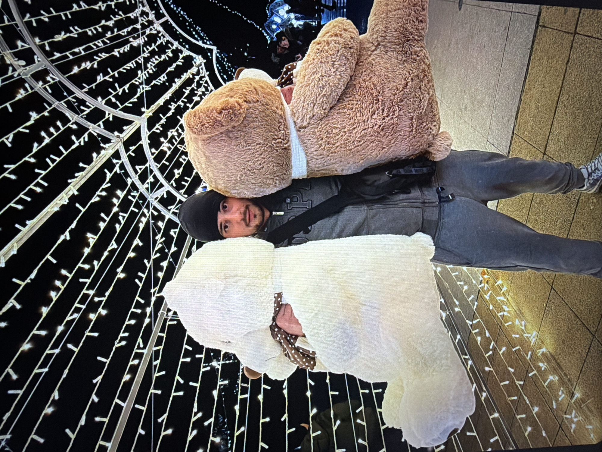 A man wearing a black beanie, gray hoodie, and gray pants is standing outdoors. A large tan teddy bear with a white bandage around its neck is draped over his shoulders. A large white teddy bear with a polka-dot bow around its neck is also draped over his shoulders. The background features a tunnel-like structure decorated with white lights, indicating a festive or holiday setting. The ground appears to be paved with light-colored tiles. The image shows details of the surrounding environment and the objects within it. While it's difficult to precisely determine the location in Burgas, Bulgaria, without additional information, the presence of festive lighting suggests a public space where seasonal decorations are common.