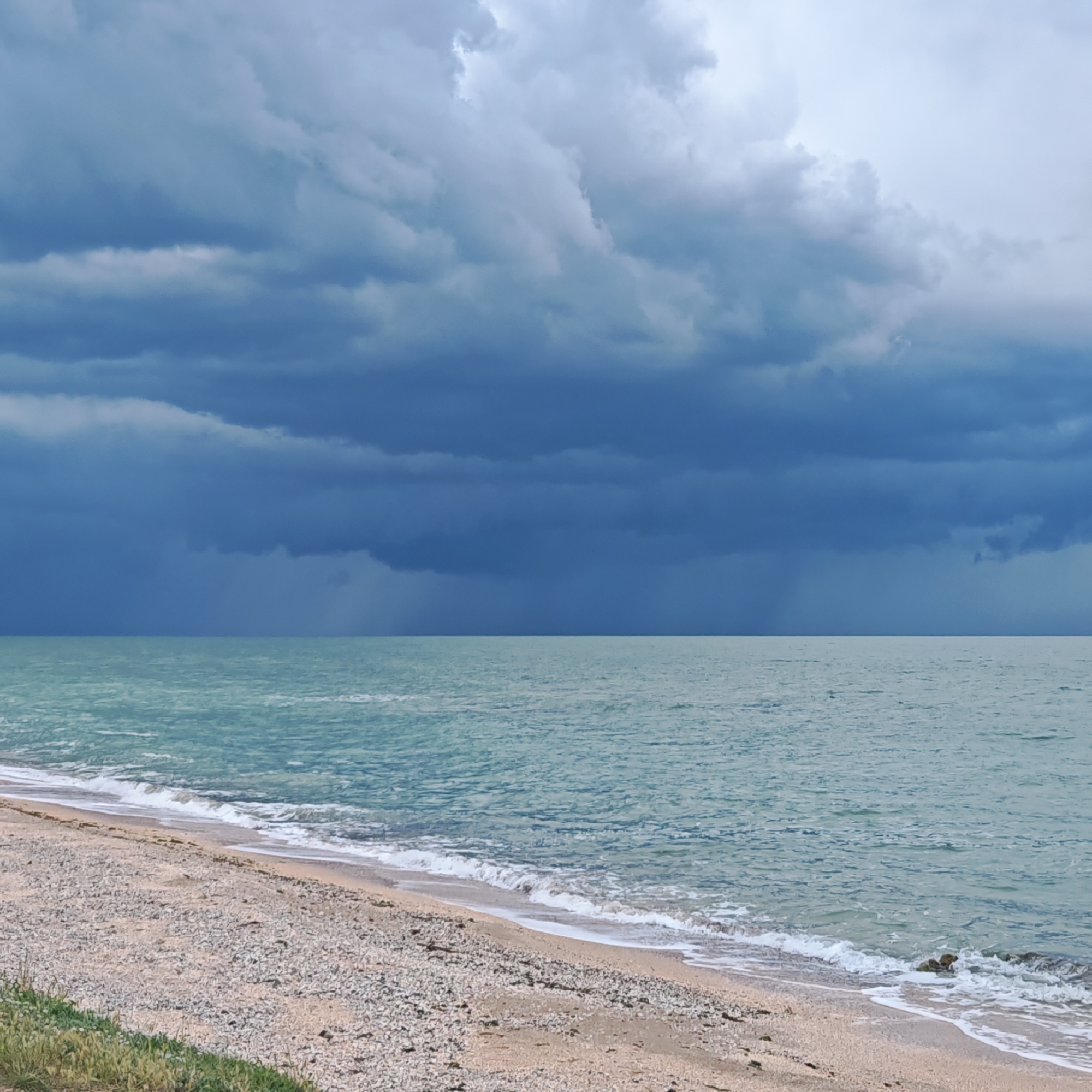 The image depicts a beach scene. Above the horizon is a band of dark, ominous storm clouds, potentially indicating a storm or rain. Below, a tranquil body of water stretches out to meet the horizon. A narrow stretch of beach is visible in the foreground, with small waves gently lapping at the shore. A small patch of green vegetation can be seen on the beach. The image does not contain any people, known locations, or interactions.