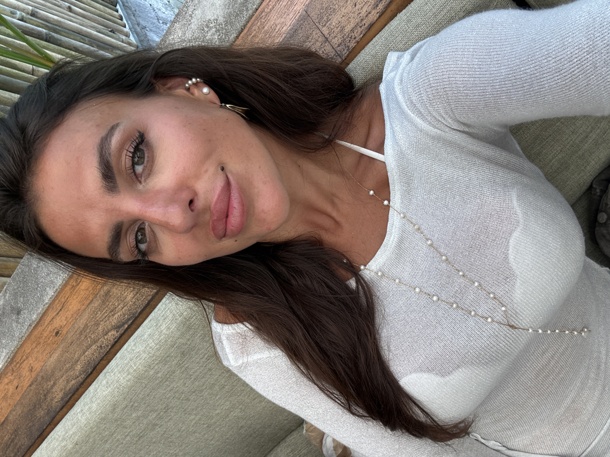 In Kuta Selatan, Indonesia, a woman with dark hair and light skin takes a selfie. She is wearing a white off-the-shoulder top and a pearl necklace. She is seated on a couch with a wooden backdrop. The woman has on light makeup and a mole on her left cheek.