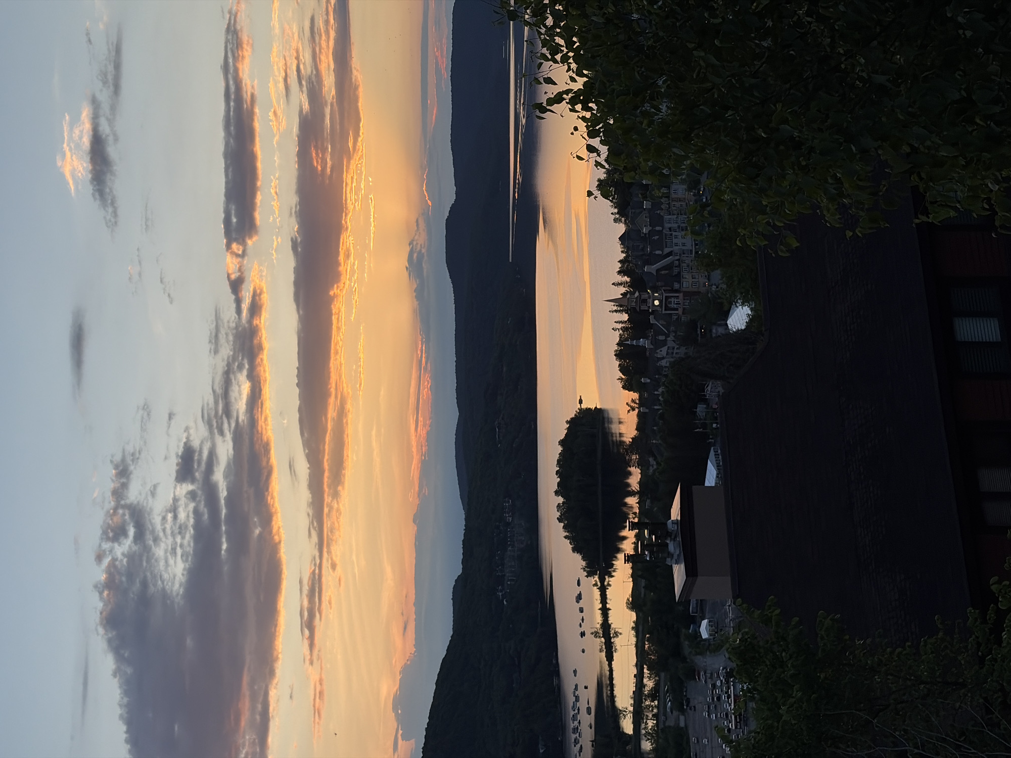 The image depicts a scenic view of Mont-Tremblant, Quebec, Canada, at dusk. The foreground is dominated by the dark roof of a building, with trees on the right partially obscuring the view. Below, a small cluster of buildings with steeply pitched roofs and a clock tower is visible, suggesting a village or resort setting. Further in the midground, a body of water, likely a lake, reflects the warm hues of the sunset. Several boats are moored on the left side of the lake, and a small, tree-covered island is situated in the water. The background features rolling, tree-covered hills silhouetted against a sky filled with a gradient of colors from pale blue at the top to vibrant orange and pink near the horizon, characteristic of a sunset. Scattered clouds add texture to the sky. The overall atmosphere is serene and picturesque, highlighting the natural beauty of the area.