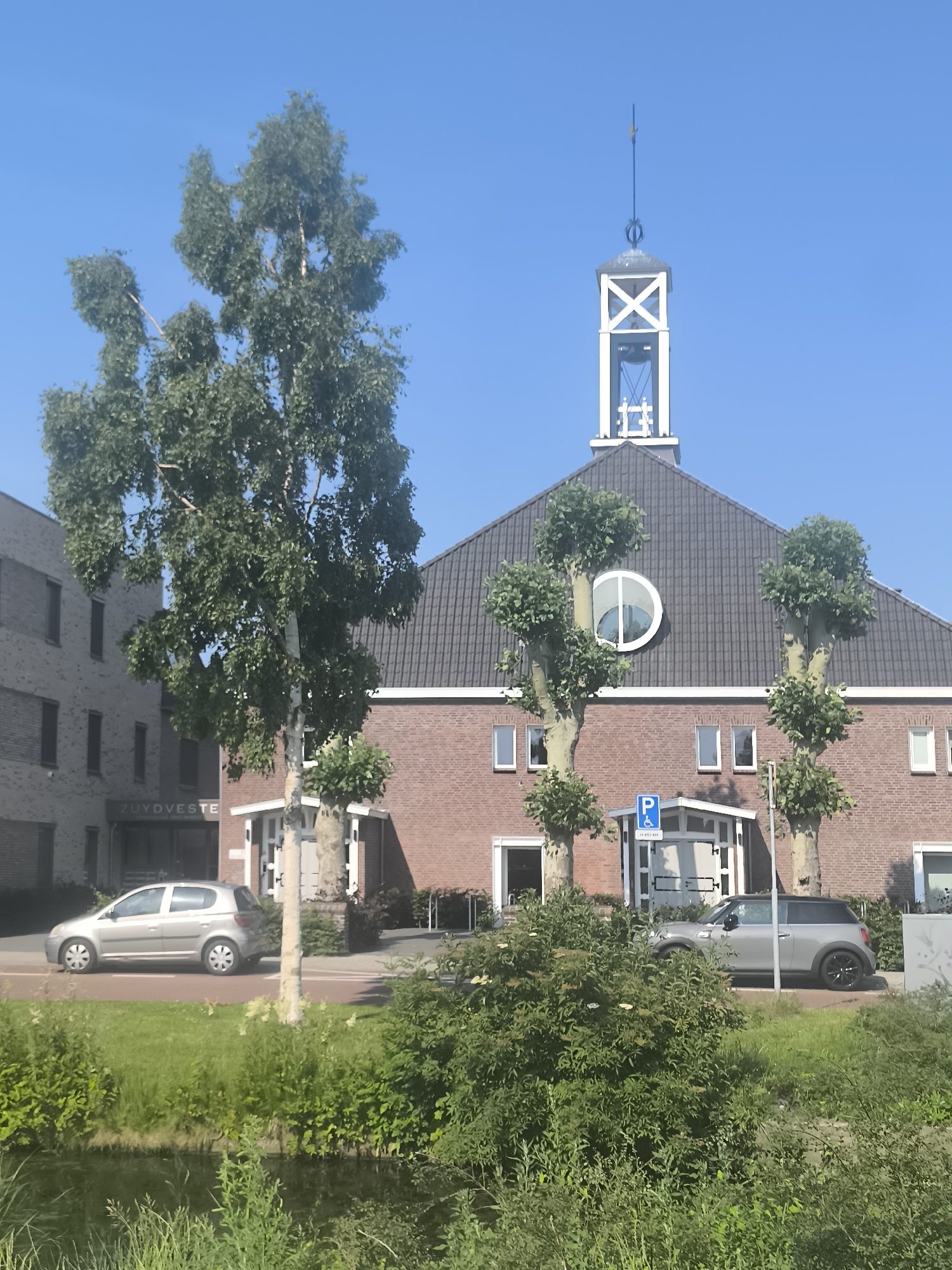 This image depicts a building with a red brick facade and a dark tiled roof, topped with a white, steeple-like structure. A circular window is visible on the facade. Two trees with distinctive, rounded canopies are planted in front of the building. To the left of the main building, a modern, multi-story brick structure is visible. Two cars are parked in front of the buildings: a silver hatchback on the left and a grey compact car on the right. A blue parking sign with a wheelchair symbol is mounted on a pole. The scene is set against a clear blue sky. The foreground features lush green vegetation and a narrow body of water, suggesting a canal or ditch. The sign on the left building reads "ZUYDVESTE". Based on the building style and the presence of the canal, this scene is likely located in Aalsmeer, Netherlands.