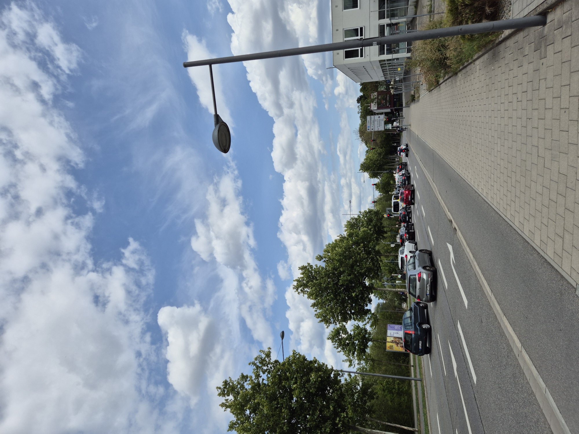 The image captures a street scene in Regensburg, Germany, on a bright, cloudy day. The view is from a sidewalk next to a multi-lane road where traffic is moving. A grey lamppost stands tall on the right side of the frame, overlooking the street and a modern, white building with large windows.
Several cars are visible on the road, including a dark blue hatchback on the left, a silver Volkswagen Golf, and other vehicles further down the road, suggesting moderate traffic. The road has clear white lane markings, including arrows indicating directions. On the left side of the road, there are trees lining the street and a billboard. The sky above is a vibrant blue with scattered white clouds, some of which are wispy and stretched across the horizon.
The sidewalk is paved with light grey rectangular bricks. In the background, more trees and buildings can be seen, indicating an urban or suburban environment. The overall impression is of an ordinary day in a German city, with traffic flowing under a partly cloudy sky.
