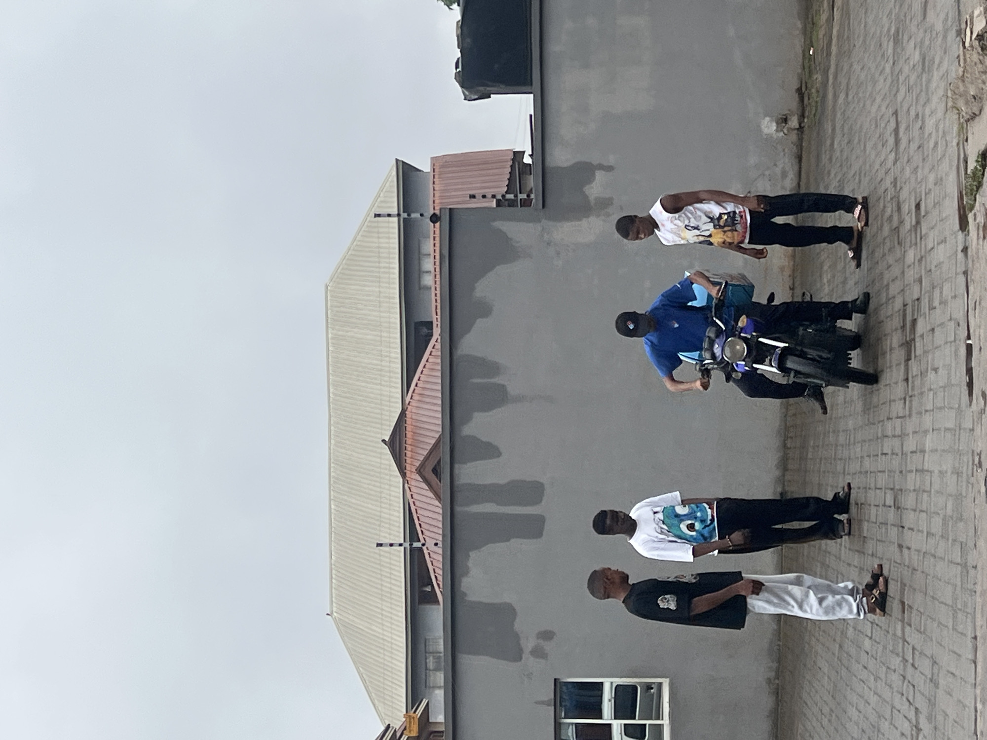 In Onitsha, Nigeria, four young men are gathered on a paved area in front of a gray concrete wall. The sky is overcast.
On the left, a man wearing a black t-shirt and white pants stands with his arms at his sides. Next to him, a man in a white t-shirt with a blue cartoon image and black pants looks down. In the center, a man in a blue t-shirt and black pants is seated on a blue and black motorcycle, holding onto the handlebars. A blue delivery box is attached to the front of the motorcycle. To the right, a man wearing a white sleeveless t-shirt with a graphic print and black pants stands with his hands clasped in front of him.
The background features a multi-story building with a light-colored, corrugated metal roof and a red-tiled roof section. A black water tank is visible on the top right of the building.