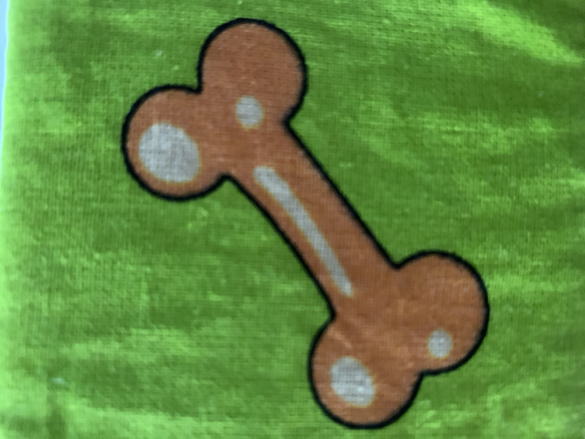 The media file is a close-up shot of a cartoon bone against a bright green, textured background, likely fabric. The bone is orange with a black outline. It has two circular, lighter colored markings on each end and a lighter, elongated marking down the center of its shaft. The green background has a subtle weave pattern visible, suggesting it's a piece of cloth or a similar textile. There are no people, scenes, or specific location cues to suggest a particular time of day, weather, or emotion. No text is visible in the image.