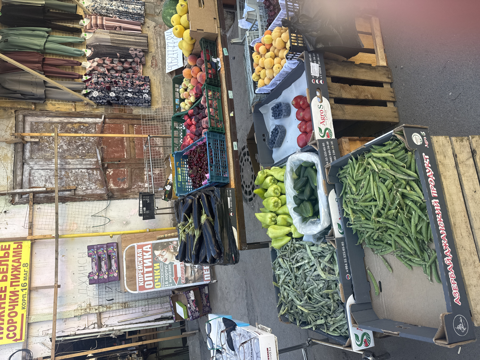 A street market stall in Saint Petersburg, Russia, displays a variety of fresh produce and clothing. In the foreground, crates of green beans, cucumbers, and green peppers are arranged on the asphalt. Behind them, a stall features eggplants, cherries, peaches, apricots, and other fruits. In the background, racks of clothing are hung on a wall, and a sign for "Korean Optics" is visible. The market appears to be outdoors, with stalls set up along a street. The overall atmosphere suggests a vibrant and busy market scene.