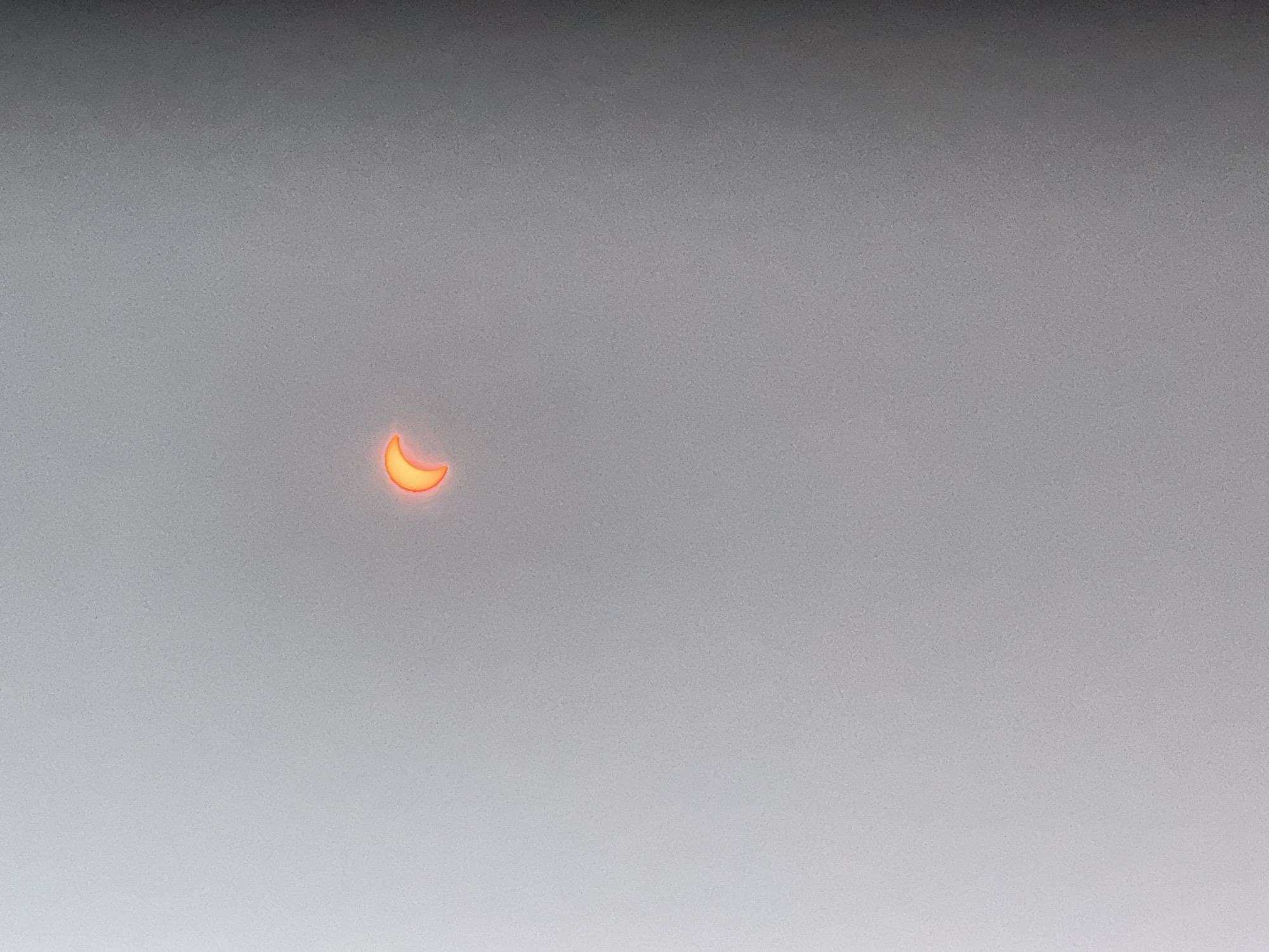 The image shows a crescent-shaped object with a bright orange glow against a muted gray background. The object appears to be a partial solar eclipse. The background is textured with a fine grain. The image is centered and the object is small in relation to the overall frame. There are no people, buildings, or other identifiable landmarks visible. The location in Märstetten, Switzerland, is not discernible from the visual information provided.