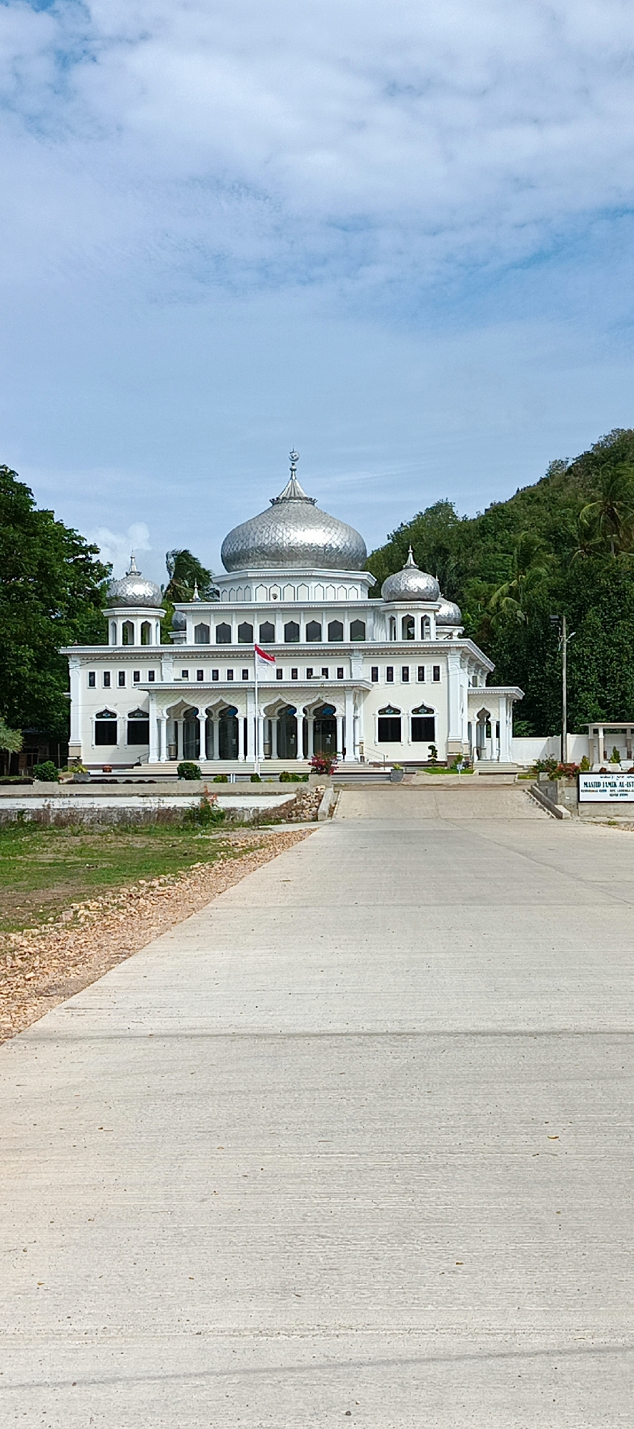 The image depicts a white mosque with multiple domes, including a large central dome and smaller domes on either side. An Indonesian flag flies in front of the mosque. The building is situated in a landscape with lush green trees and a hill in the background, under a partly cloudy blue sky. A paved road leads up to the mosque, with a gravel border on the left side. A sign is visible on the right side of the road in front of the mosque, which appears to read "MASJID JAMIK AL-AST...". The scene is an exterior view of a public building in a tropical or subtropical environment, likely located in the city of Tanjong, Indonesia, given the presence of the Indonesian flag and the architectural style which is consistent with the region.