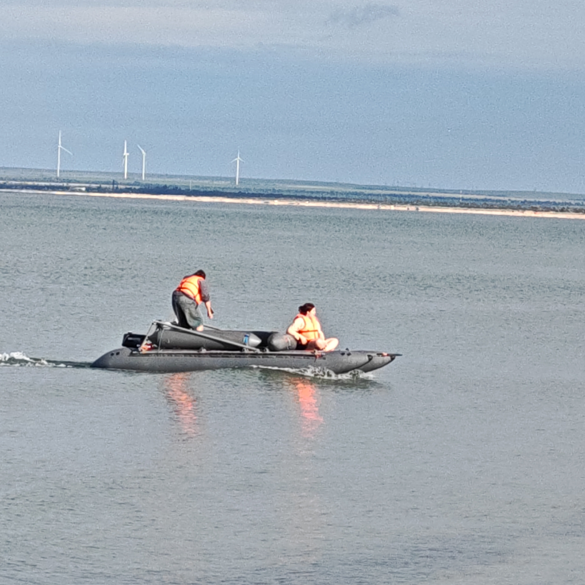The image shows a small, dark-colored inflatable boat moving on a body of water. Two people are on board. One person is standing towards the back of the boat wearing an orange life vest, and the other is sitting cross-legged near the front of the boat, also wearing an orange life vest. The boat appears to be creating a wake as it moves through the water. In the background, there is a strip of land with a few wind turbines visible on the horizon. The sky is overcast. The location is Unknown, Unknown.