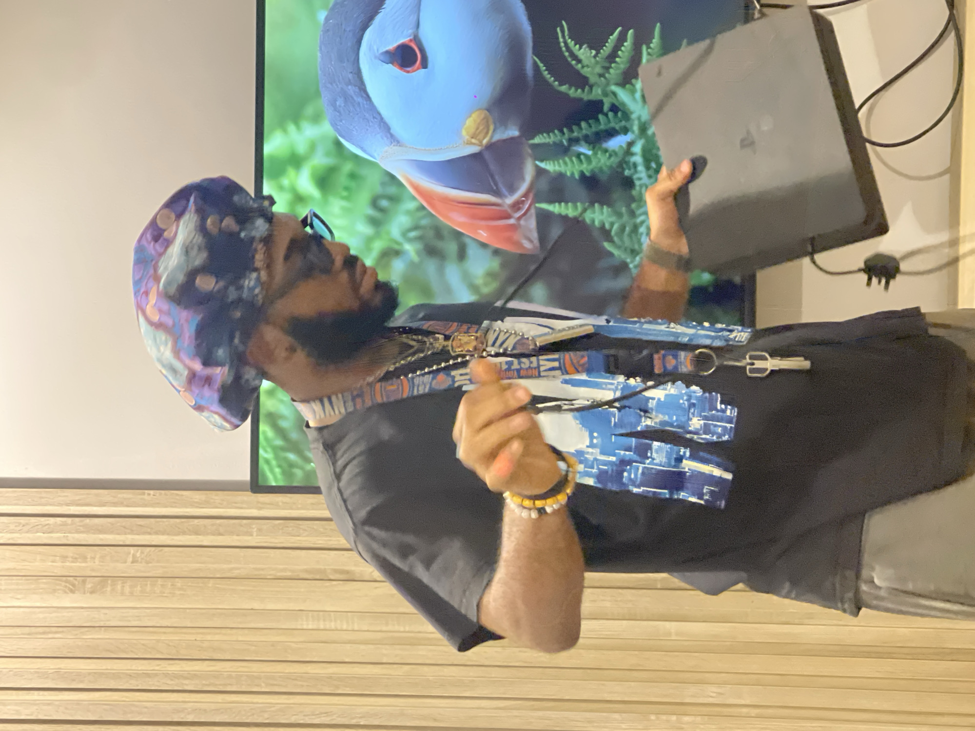 The image shows a man wearing a black t-shirt and a patterned headscarf. He has a beard and is wearing glasses. He is holding a dark grey electronic device, possibly a game console, in his right hand. He is standing in front of a large screen displaying an image of a puffin. The man is wearing a lanyard around his neck with several keychains attached. He also has bracelets on his wrist. The background shows a light-colored wall and wooden paneling, suggesting an indoor setting. The image is brightly lit, and there are no clear indications of the time of day or weather. The man's expression is difficult to discern. The scene is likely in a home or entertainment space.