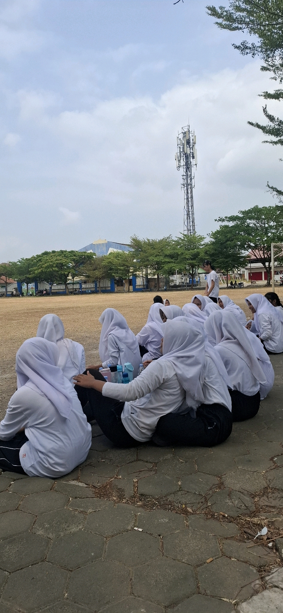 In Baturan, Indonesia, a group of individuals, predominantly young women wearing white hijabs and uniforms, are seen sitting on a paved hexagonal surface outdoors. The group appears to be engaged in a break or activity, with some individuals resting and others interacting. In the background, a tall telecommunications tower stands prominently against a cloudy sky. Adjacent to the tower, there are trees and a blue-and-white building, possibly a school or sports facility. The ground is a mixture of dry, tan-colored soil and the hexagonal pavement. Scattered debris, including dry leaves and cigarette butts, is visible on the pavement. A person in a white t-shirt and dark pants stands in the background, observing the group. Several water bottles are placed on the ground among the seated individuals. The overall scene suggests an outdoor gathering or activity in an urban or semi-urban environment.