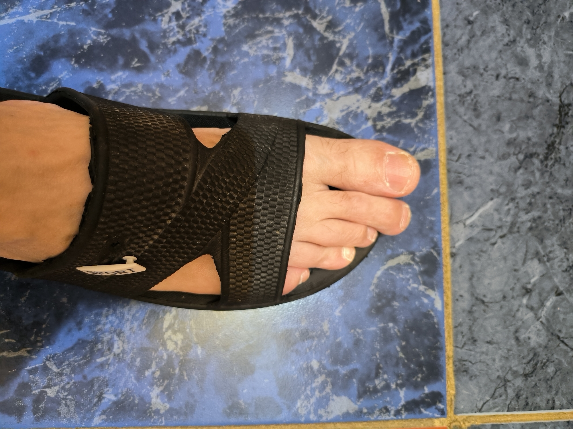 A person's foot, wearing a black slide sandal, rests on a blue and grey tiled floor. The sandal has a textured strap and a small white tab with the word "SPORT" embossed on it. The tiles have a marbled pattern. The scene is indoors, and it is impossible to determine the specific location within Yuzhno-Sakhalinsk, Russia from the image alone.