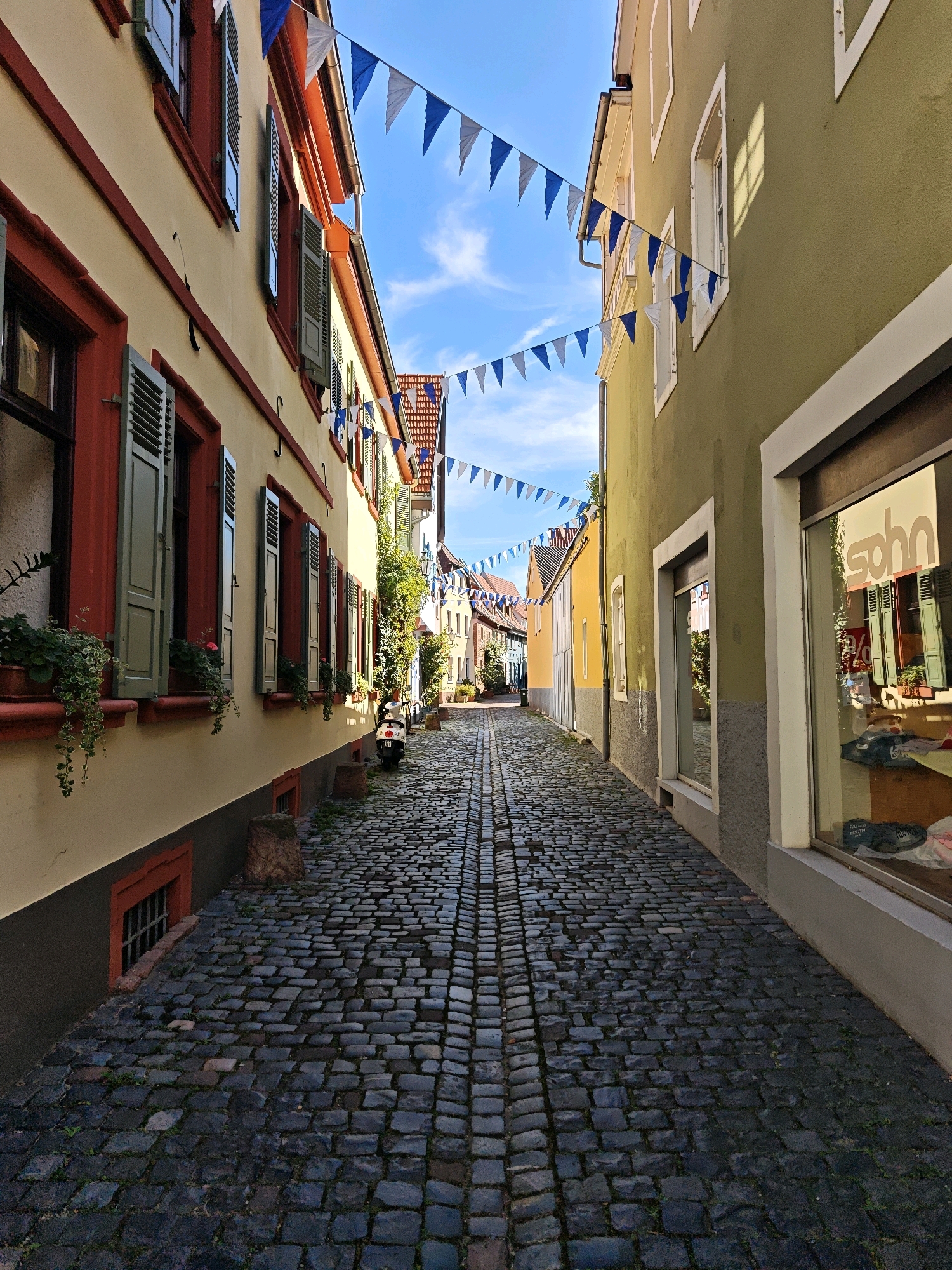 The image depicts a narrow cobblestone street in the historic city of Ladenburg, Germany, lined with colorful buildings adorned with festive triangular flags. The street is paved with dark, uneven cobblestones, with patches of green growth emerging between them. On the left, a building with a pale yellow facade features red trim around windows and shutters painted in a muted green. Planters with hanging greenery and small flowers are placed on the window sills. Further down the street on the left, a white scooter is parked.
The building on the right has a pale green facade with white trim around its windows. One large storefront window on the right reflects the building opposite and signage that reads "sohn". Inside the window, clothing and mannequins are visible.
In the background, the street recedes, with more buildings visible on either side, some with red-tiled roofs. The sky above is a bright blue with scattered white clouds. The overall impression is of a charming, old European street scene, likely decorated for a local festival or event.