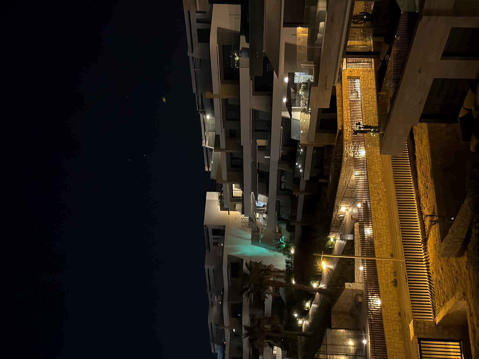 The image depicts a nighttime view of a modern residential complex located in Finestrat, Spain. The architecture features white, multi-level buildings with balconies and large windows. Some buildings are illuminated with warm yellow lights, while others have a striking green light emanating from their facades, creating a dramatic effect against the dark sky. Palm trees are visible in the foreground, adding to the Mediterranean atmosphere. The ground level shows illuminated pathways and stone walls, with vertical fences casting long shadows. The overall scene conveys a sense of upscale living and tranquil evening ambiance in a coastal Spanish town.