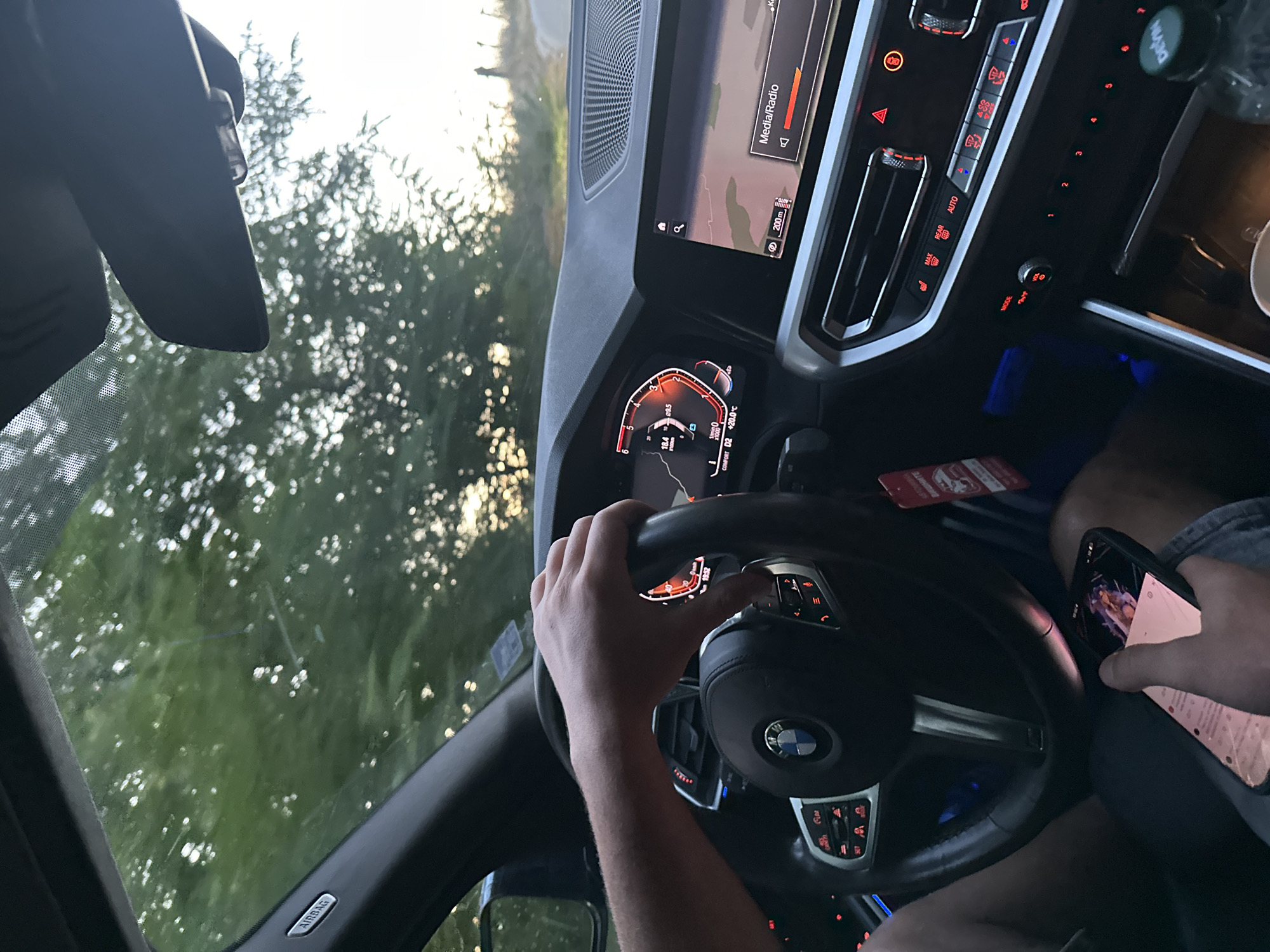The image shows a person's hands inside a car, likely a BMW, based on the steering wheel emblem. One hand is on the steering wheel, while the other is holding a smartphone. The dashboard displays information on a digital screen, including navigation and media options, with the word "Media/Radio" visible. The car's interior is dimly lit, with some ambient blue light emanating from beneath the dashboard. Outside the car, trees and a sunset are visible through the windshield, suggesting the car is in a rural or semi-rural setting. The photograph was taken in Kozarka, Bulgaria, indicating a specific location. The overall scene depicts a moment of driving, with a focus on the car's interior and the driver's interaction with their phone.
