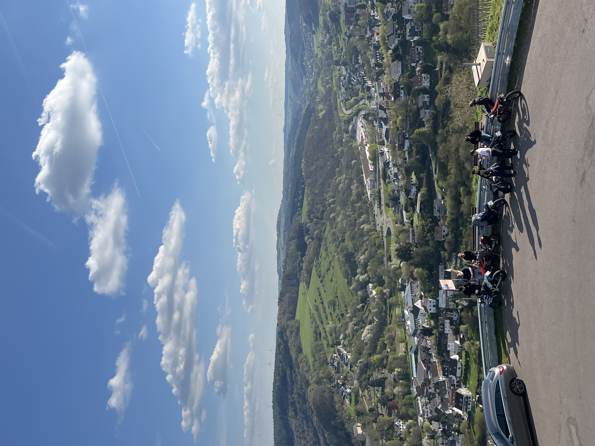 The image depicts a scenic overlook point in Saarburg, Germany. Six individuals are positioned along a guardrail with motorcycles parked beside them, suggesting a break during a ride. The vantage point offers a panoramic view of the town below, characterized by closely packed residential buildings amidst lush greenery. The landscape extends into rolling hills and forests, with wind turbines visible in the distance. A clear blue sky with scattered cumulus clouds and contrails provides a bright background. A car is parked at the edge of the road nearby, suggesting vehicular access to the viewpoint.