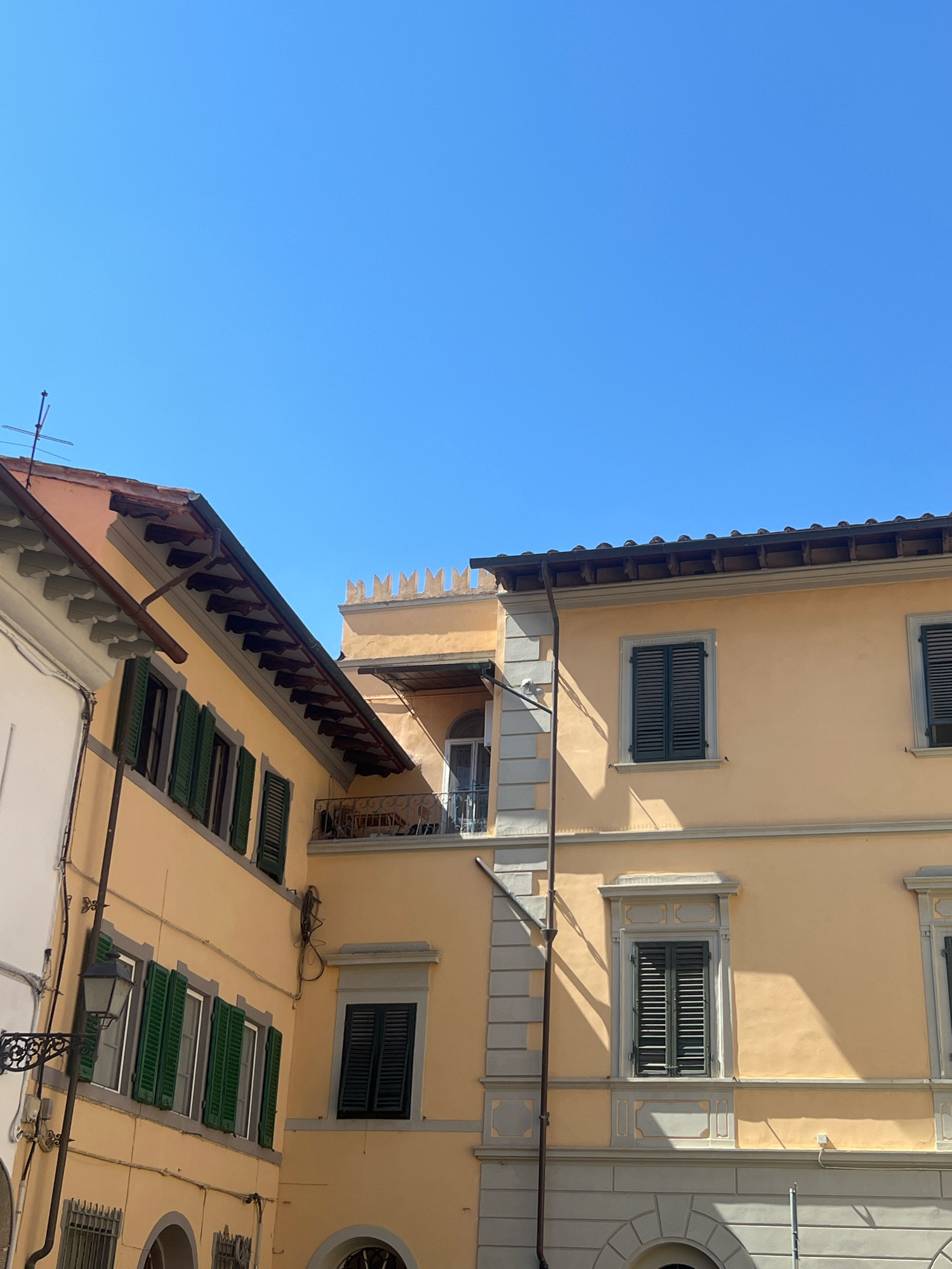 The image shows an exterior view of two adjacent buildings with pale yellow facades under a clear blue sky. The building on the left is angled and features a terracotta tiled roof with exposed wooden eaves. It has multiple windows with green shutters, some of which are open. A dark gutter runs along the roofline, and a rustic black lantern hangs on the left side of the building. The building on the right is taller and has a more ornate architectural style with decorative grey stone detailing around the windows and at the top. It also features windows with dark shutters and a balcony with a wrought-iron railing. The sun casts shadows across the facades, indicating a bright, sunny day. The scene appears to be in a city, possibly a historic area, with elements suggesting a European architectural style. There are no people visible in the image.