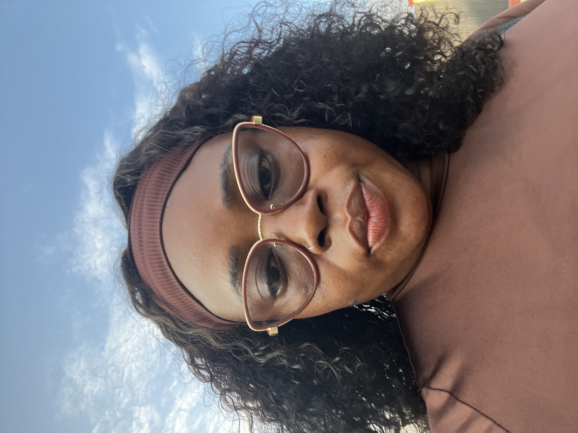 A close-up, low-angle selfie of a Black woman with dark, curly hair, wearing a brown headband and oversized cat-eye glasses. She is also wearing a brown shirt. The background shows a bright blue sky with scattered white clouds. The overall scene suggests an outdoor setting, possibly in the city of Abuja, Nigeria, given the natural lighting and clear sky. No specific city landmarks are visible to confirm the location.