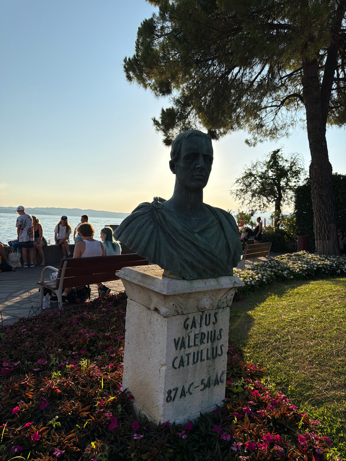 The image shows a bronze bust of Gaius Valerius Catullus, a Roman lyric poet, placed on a stone pedestal. The inscription on the pedestal reads "Gaius Valerius Catullus 87 BC - 54 BC". The bust is positioned in a park-like setting with a large tree providing shade, and a lawn with flowers in the foreground. In the background, there are people sitting on benches near a body of water, possibly Lake Garda. This is likely located at the Grotte di Catullo in Sirmione, Italy, an archaeological site featuring the ruins of a Roman villa and a museum dedicated to the poet Catullus. The sun is setting, casting a warm glow on the scene.