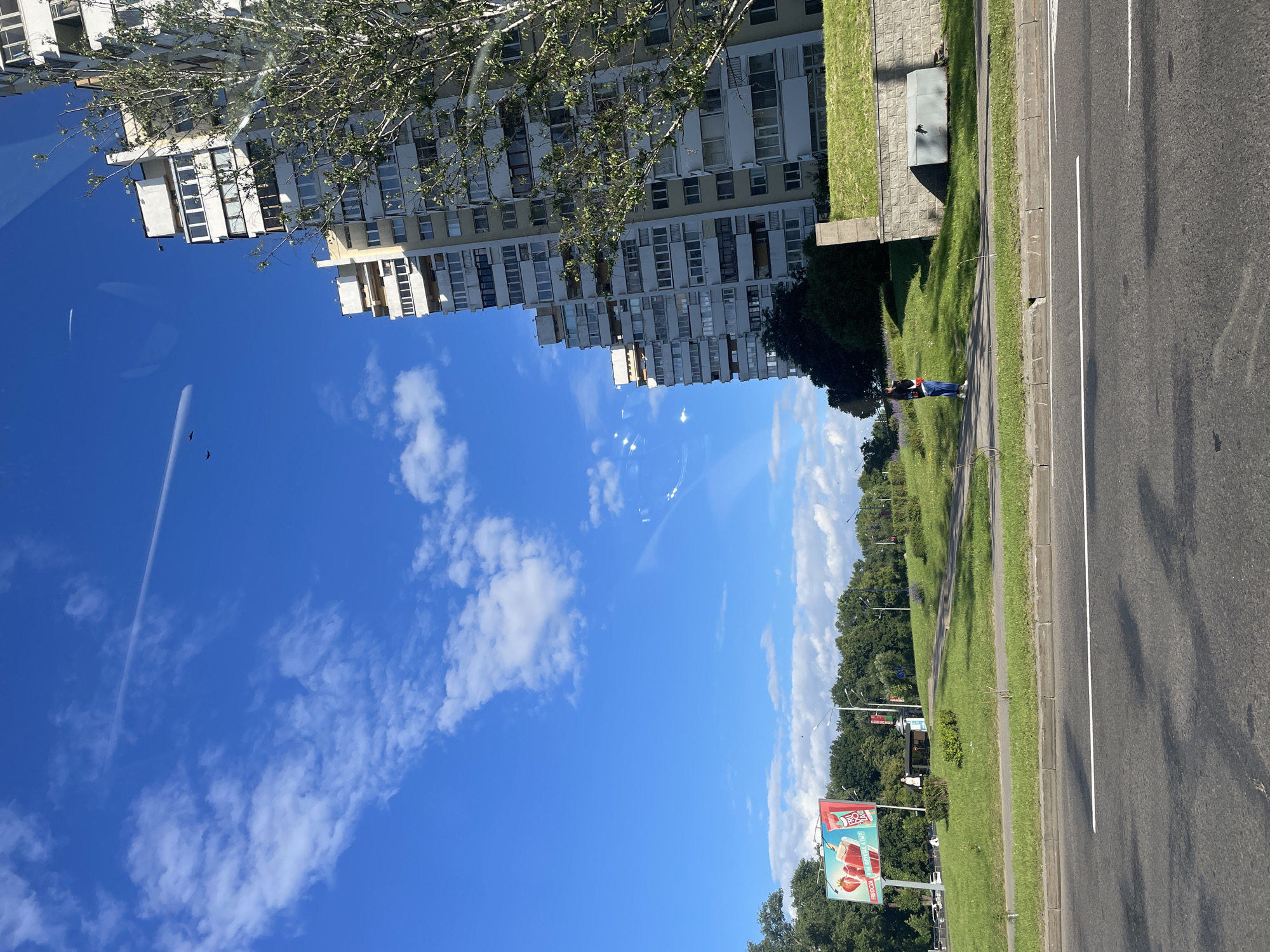 The image depicts a sunny day with a bright blue sky and scattered white clouds. A tall, multi-story apartment building dominates the right side of the frame, its balconies and numerous windows visible. A tree with sparse foliage stands in front of the building. In the background, a billboard advertising "FRUOCZA" with images of strawberries and a drink is visible, along with a bus stop shelter and lush green trees. A paved road occupies the foreground, with a sidewalk and grassy embankment between the road and the buildings. A single person is standing on the sidewalk, looking down at their phone. The overall scene suggests an urban environment in Minsk, Belarus, likely during daytime.