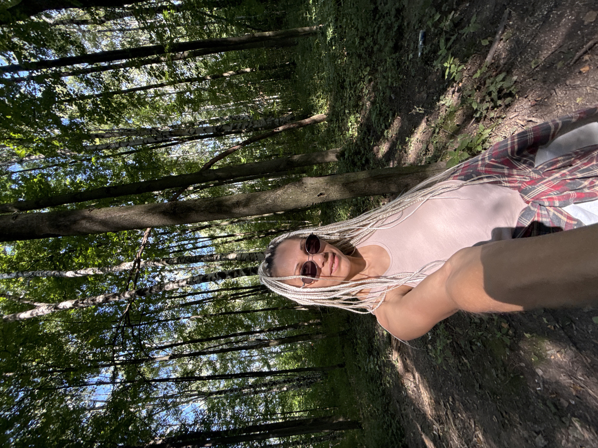 The image is a selfie taken by a woman with blonde braided hair in a forest in the city of Rusanovo, Russia. She is wearing sunglasses, a pink tank top, and a red and black plaid shirt tied around her waist. The background is a dense forest with tall trees and green undergrowth. The sun is shining through the trees, creating dappled light on the ground. The woman is smiling and looking at the camera.