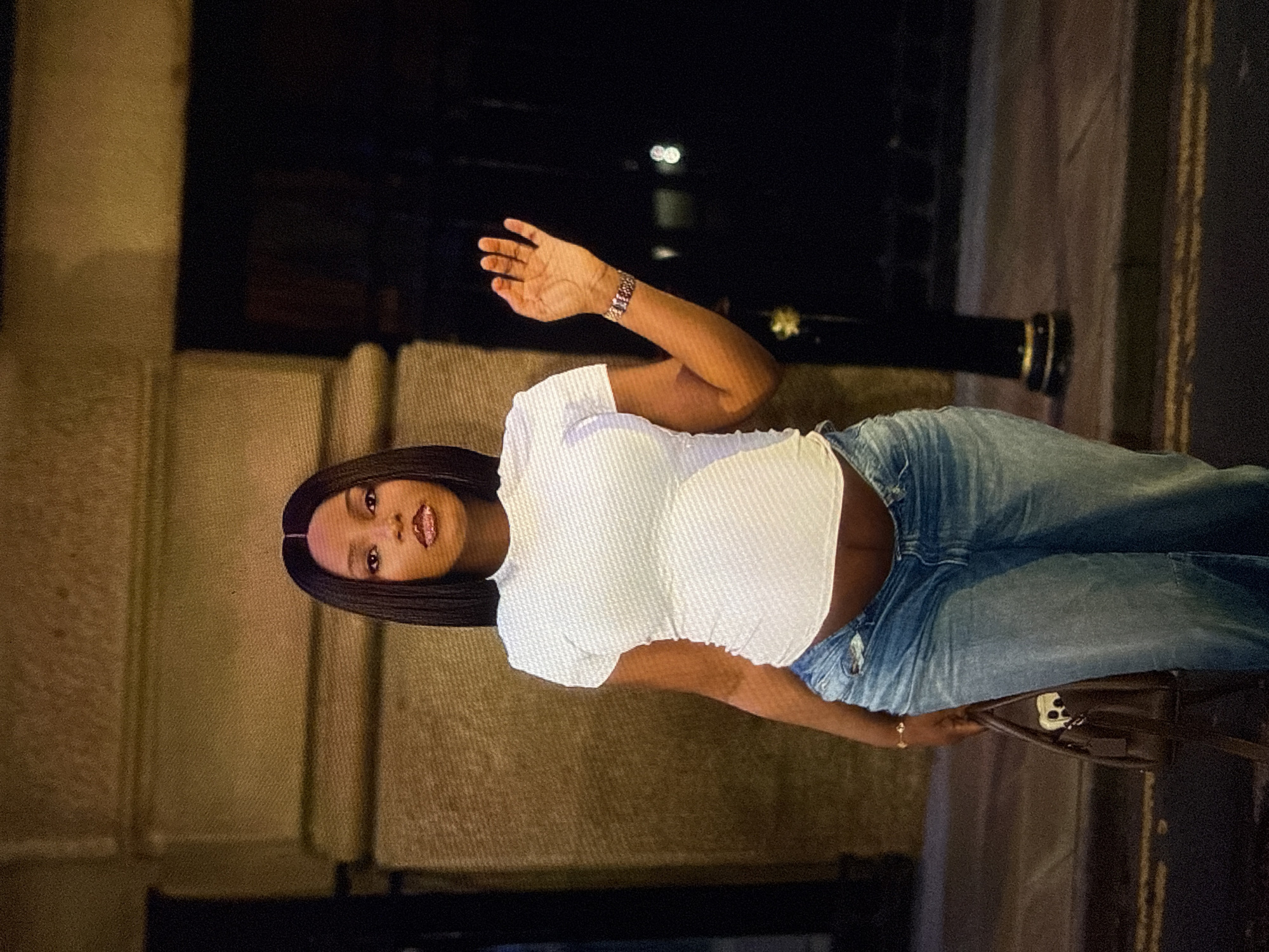 A Black woman stands in front of a building with textured walls, possibly at dusk in an urban setting. She has straight, dark hair parted in the middle and is wearing a white, short-sleeved, form-fitting shirt that exposes her midriff, and light-wash, baggy jeans. Her left hand is raised, palm facing forward, and she wears a watch on her left wrist. She is holding a dark brown handbag in her left hand. The background is dimly lit, with a dark, indistinct building to her right and a lamppost. The ground appears to be a paved surface with yellow road markings. The lighting suggests an outdoor environment at night or in low light conditions.