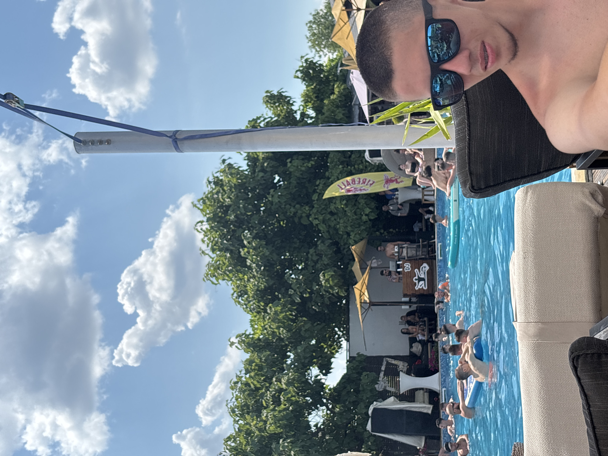 The image depicts a sunny day scene at a poolside venue, possibly a club or resort, in Krasen, Bulgaria. The foreground is dominated by a young man wearing sunglasses and looking towards the camera. He is seated in a dark chair, with part of a light-colored chair visible to his left. Behind him, a vibrant blue swimming pool is filled with people. Some individuals are swimming or lounging on inflatable floats, while others are gathered near a DJ booth situated in the background. A yellow flag with red lettering is visible near the DJ booth. Tall green trees form a backdrop behind the pool area, and a bright blue sky with scattered white clouds is visible above. The overall atmosphere suggests a social gathering with music and leisure activities.