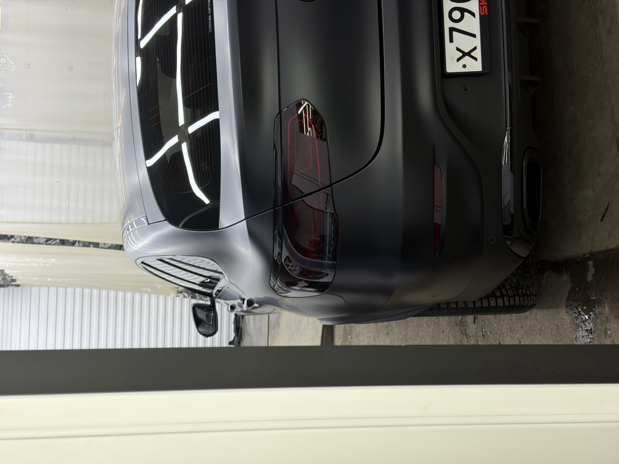 The image shows the rear of a matte black Mercedes-Benz GLC-Class SUV, license plate number *.X790 SK*, parked in what appears to be a garage or vehicle detailing space. The SUV is angled slightly towards the left of the frame. The rear window has grid-like reflective patterns, and the taillights are smoked. A tire is visible on the lower right, positioned on a concrete surface with some moisture or liquid present. A white or light-colored wall or barrier is in the foreground, partially obstructing the lower portion of the view. The location is Krasnoyarsk, Russia, due to the Russian license plate.