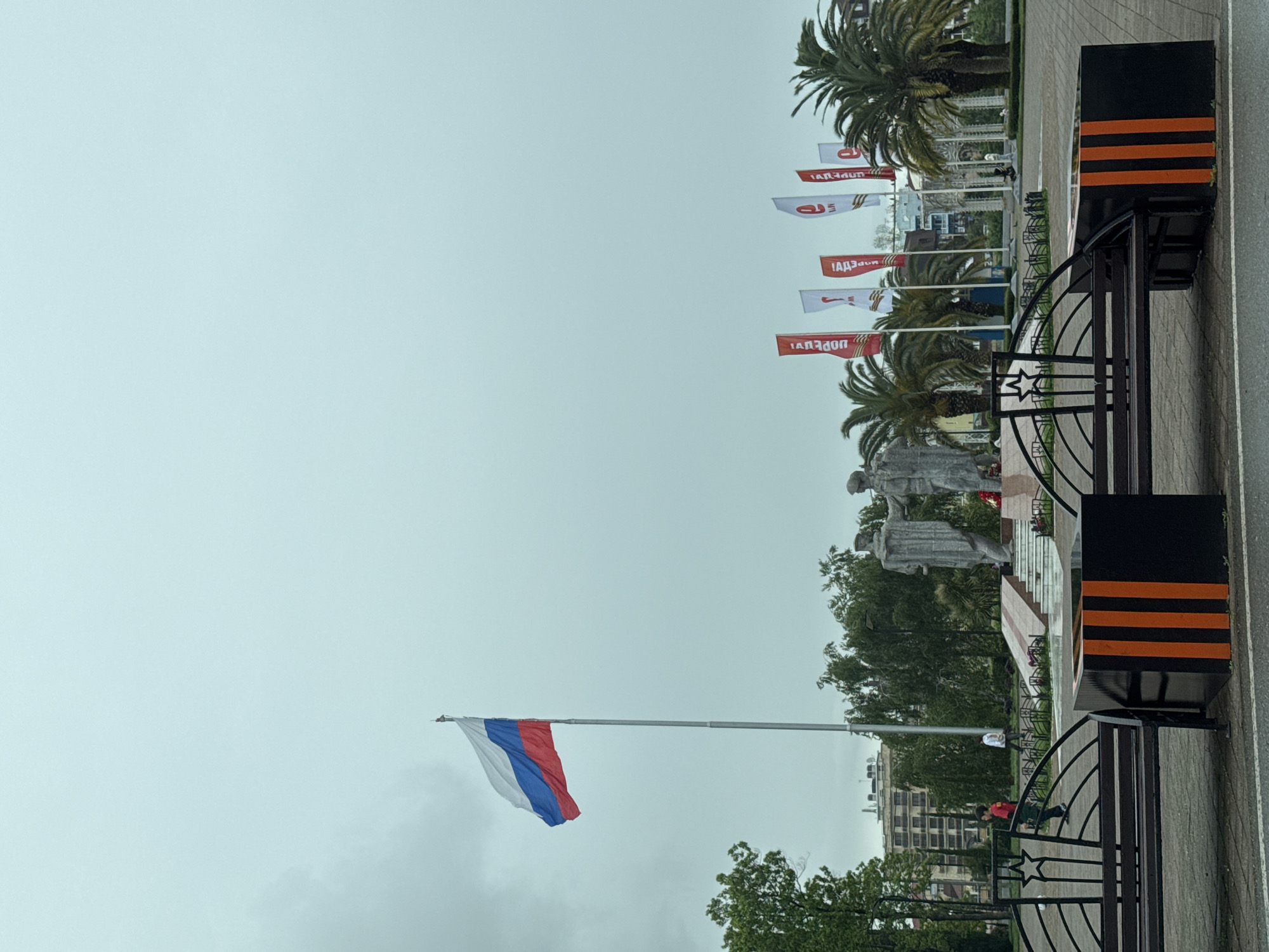 The image depicts a Russian flag waving in the sky against a cloudy backdrop in Sochi, Russia. In the background, there's a monument with statues and a series of flags with red and white coloring. The scene also includes palm trees, buildings, and decorative elements, such as black and orange striped planters with metal fences featuring star details. There are people moving in the background.