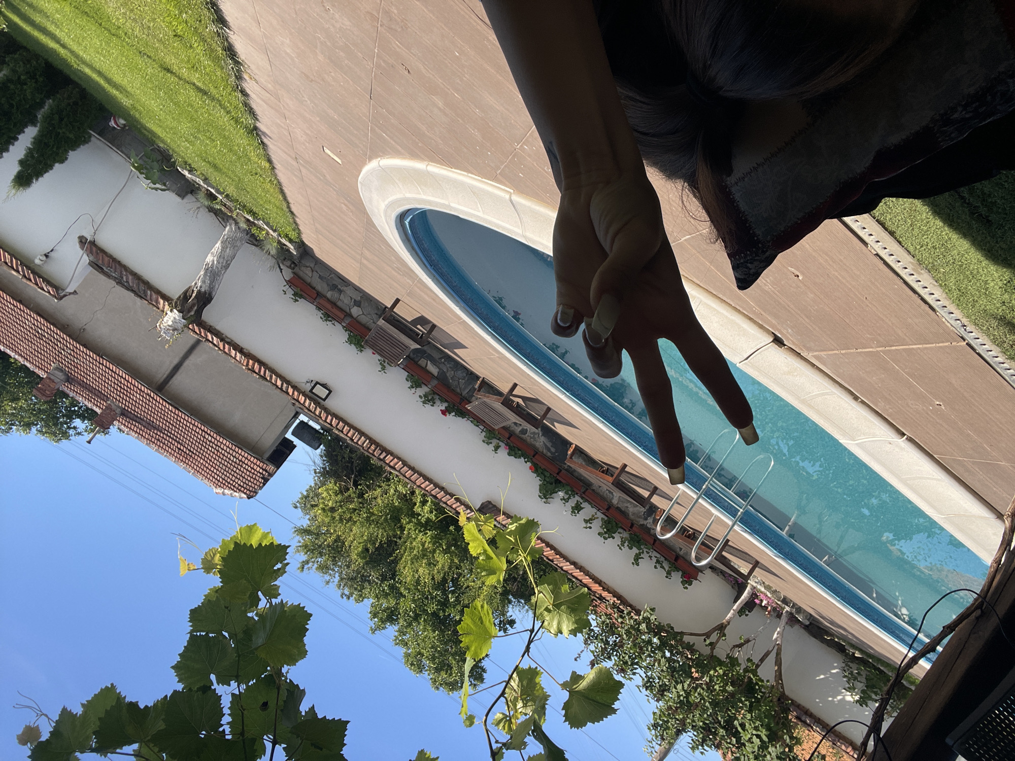 A first-person perspective shot shows a hand making a peace sign in front of a swimming pool in a backyard. The hand has long, light-colored nails with a metallic sheen. The swimming pool is oval-shaped with bright blue water and a white edge. The pool area is paved with light brown tiles, and there is a strip of green grass to the left of the pool. In the background, a white wall runs along the property line, with red-tiled roofs of houses visible above it. There are trees and some lounge chairs on the other side of the pool. The sky is clear and blue. The image appears to be taken in Totleben, Bulgaria, based on the reference to the city.
