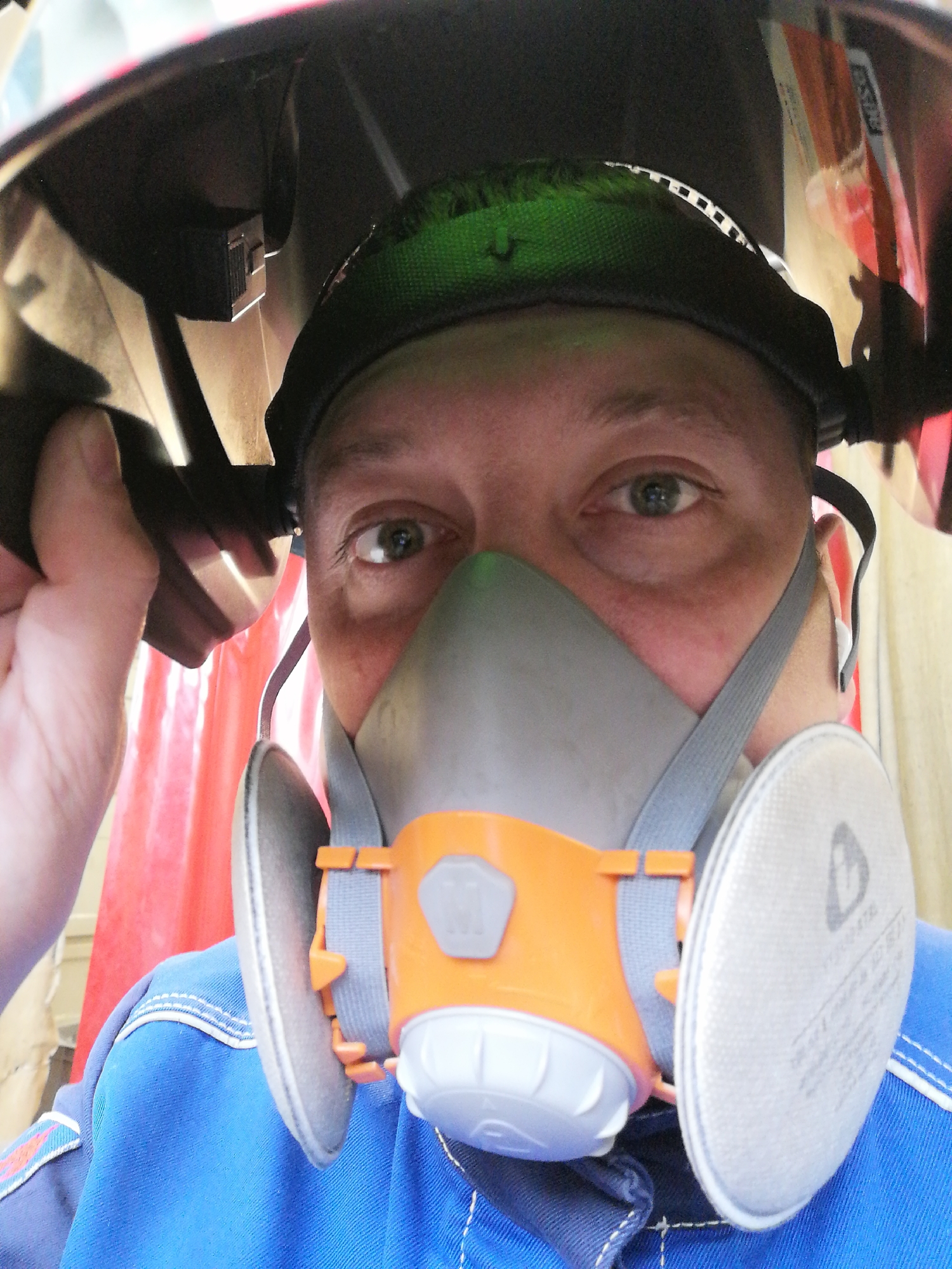 The image captures a close-up, eye-level perspective of a person, likely a man, adorned in industrial safety gear. His face is largely obscured by a grey and orange half-facepiece respirator mask, which covers his nose and mouth, but his focused eyes are visible. He is wearing a blue work overall with distinct white stitching.
The person's right hand is holding up a dark welding helmet, which is partially lifted, revealing the internal structure and a green padded head-liner. This action suggests he is either preparing to engage in or has just finished a task requiring welding or similar heavy-duty protection.
In the background, a vibrant red, fabric-like material, possibly a welding curtain or partition, is visible on the left, contrasting with lighter, indistinct surfaces on the right.
Notable details include clear branding and certification information on the respirator mask's right filter. Text visible includes "3M", "6035", and "P3 R", along with the standard "EN143:2000", identifying it as a high-efficiency particulate filter. The brand "ESAB" is also legible on the green headwear inside the welding helmet. The overall scene conveys a professional and safety-conscious work environment.