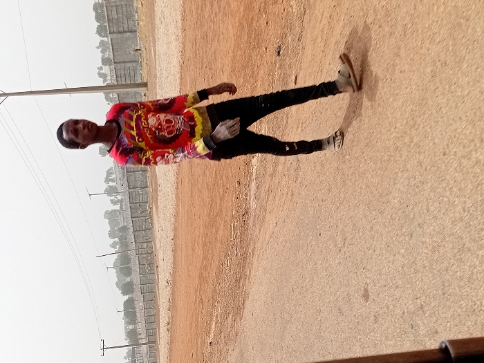 The image displays a male subject walking on a paved surface in Katcha, Nigeria. The subject, appearing to be a young adult, possesses a dark complexion and short, dark hair. He is attired in a long-sleeved shirt featuring a multi-colored tie-dye pattern primarily in red, yellow, black, and purple, with a graphic design on the chest. He wears black trousers and light-colored sandals. The subject is oriented with his body angled slightly towards the right side of the frame, moving generally leftward. To the subject's right is an unpaved area of reddish-brown earth. The background features a low concrete block wall or fence spanning horizontally, behind which utility poles with overhead lines and sparse trees are visible. The sky is hazy or overcast.