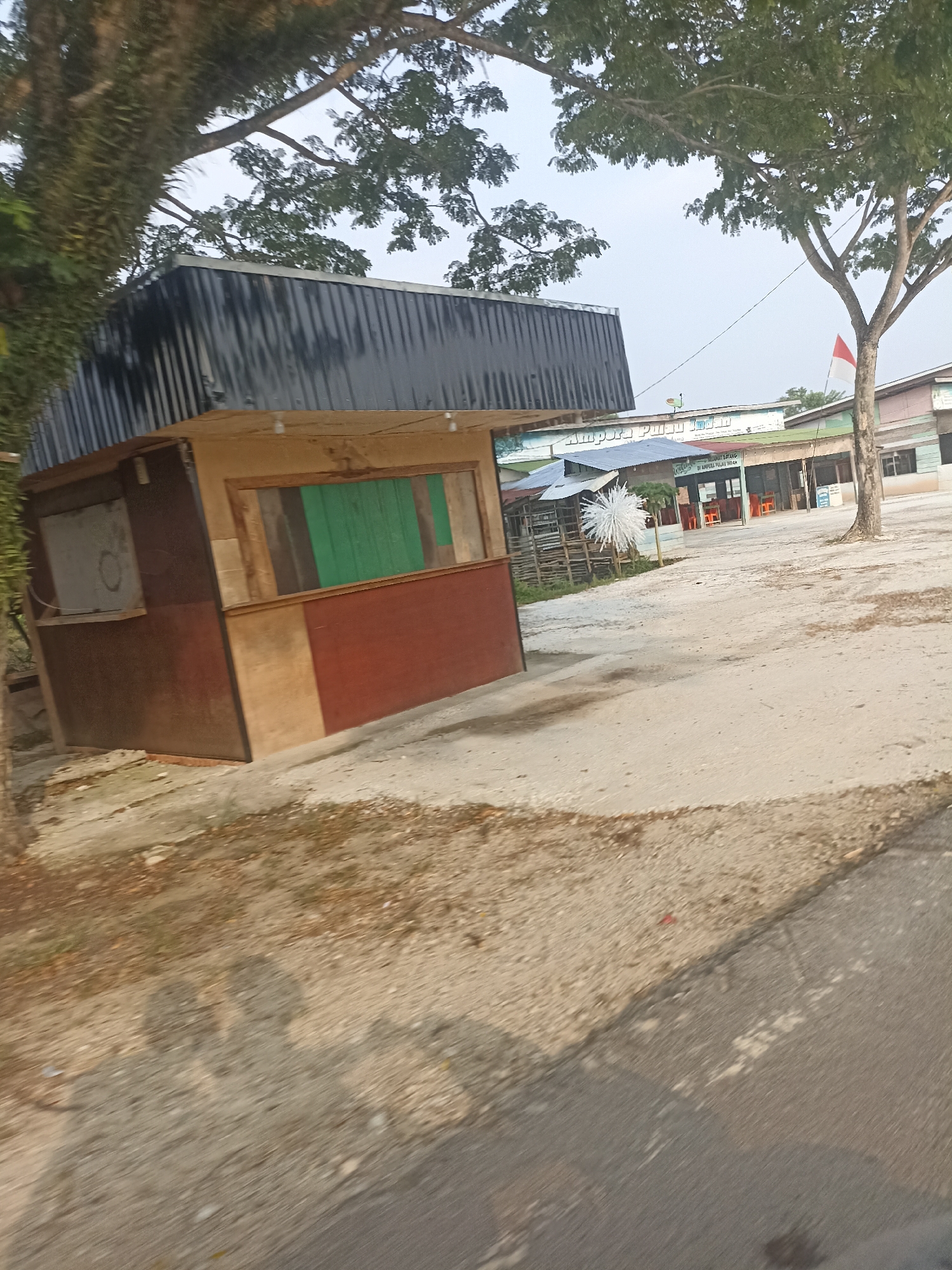 The image captures a sunlit outdoor scene in Kampung Tengah, Indonesia. In the foreground, a small, dilapidated wooden kiosk with a corrugated metal roof stands at an angle. The kiosk has a brown and red exterior, with wooden frames around two openings. One opening shows a green panel, while the other appears to be a blank board. The ground around the kiosk is sandy and dirt-covered, with some dry leaves and debris. In the mid-ground, a building complex with several structures is visible. One building features a sign with Indonesian text that appears to read "Selamat Datang Di Ampuara Pulau Merah" which translates to "Welcome to Ampuara Red Island". Another building has a red and white Indonesian flag flying. There are orange chairs visible outside some of the buildings. The environment is somewhat arid, with a large tree in the mid-ground to the right of the buildings. The sky is overcast and light. In the foreground, shadows of two people are cast on the ground, suggesting they are the photographers. The overall impression is of a roadside establishment in a developing or rural area.