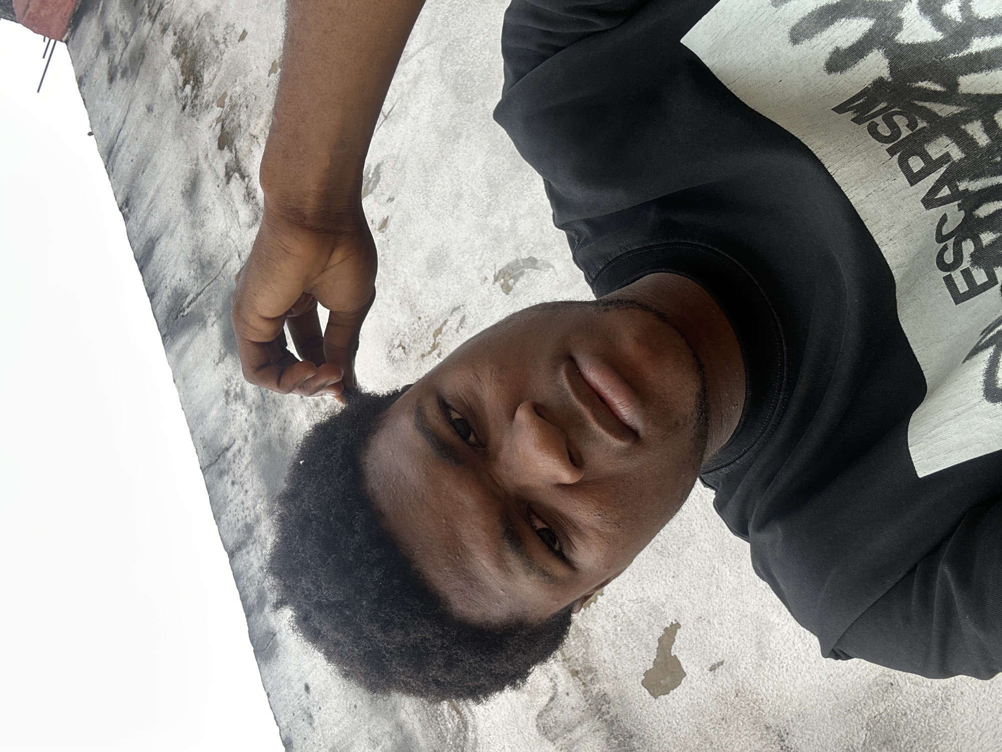 A young man with dark, curly hair is shown in a close-up, high-angle shot, looking towards the camera with a serious expression. He is wearing a black t-shirt with white lettering that appears to say "ESCAPISM." His right hand is raised, touching the side of his head. He is outdoors, with a textured, weathered concrete wall behind him and a white, overcast sky above. The environment suggests an urban or semi-urban setting.