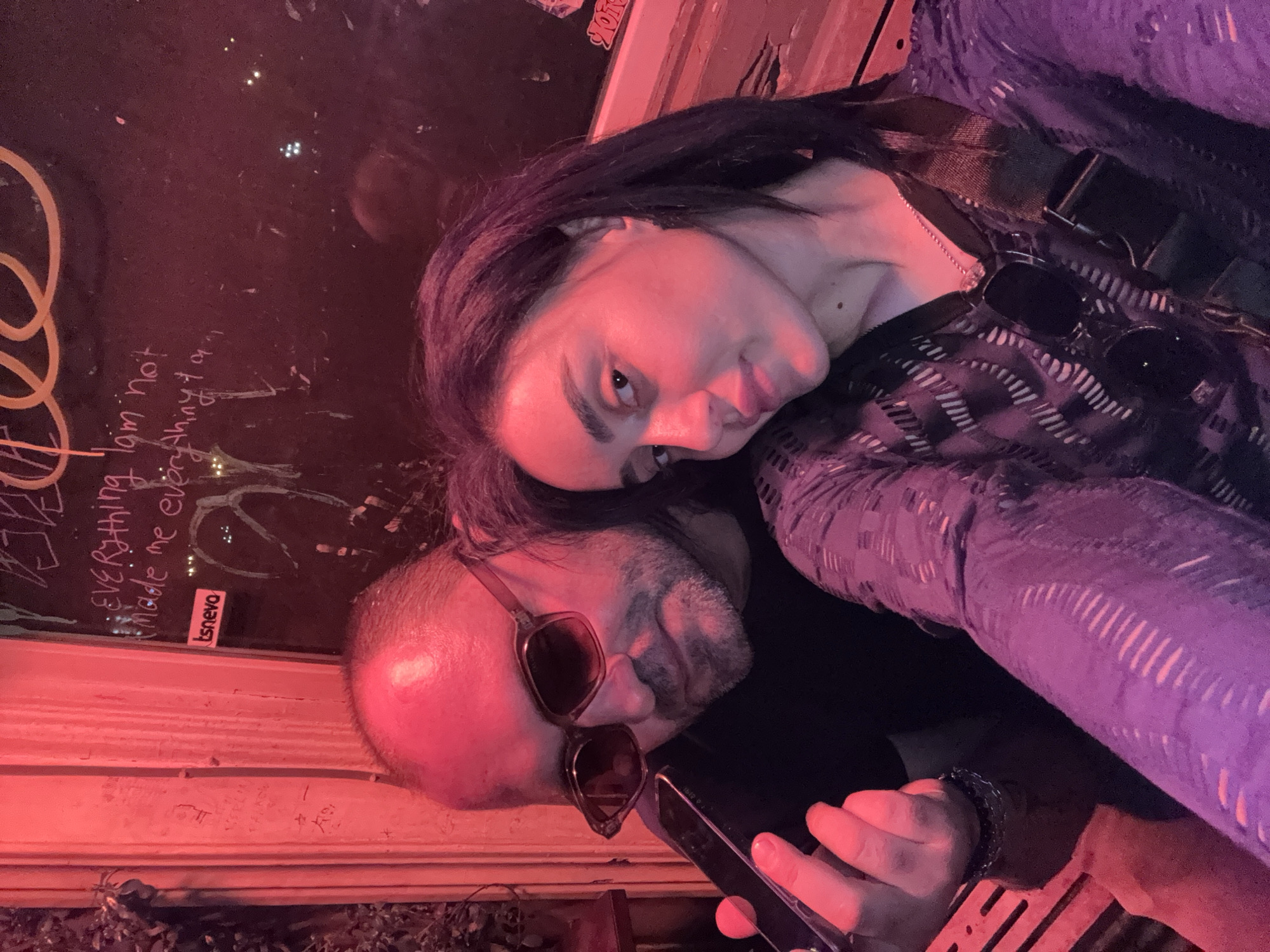 A photograph depicts a man and a woman posing closely together in an indoor or semi-outdoor setting. The location is inferred to be Tbilisi, Georgia, based on the "tsnevo" sticker visible in the photo, likely a reference to a Georgian brand or place. The woman has long dark hair and is wearing a patterned top. The man is bald, wearing sunglasses, and holding a mobile phone in his hand. The light in the scene is red. Behind the couple is a glass surface with writing on it.