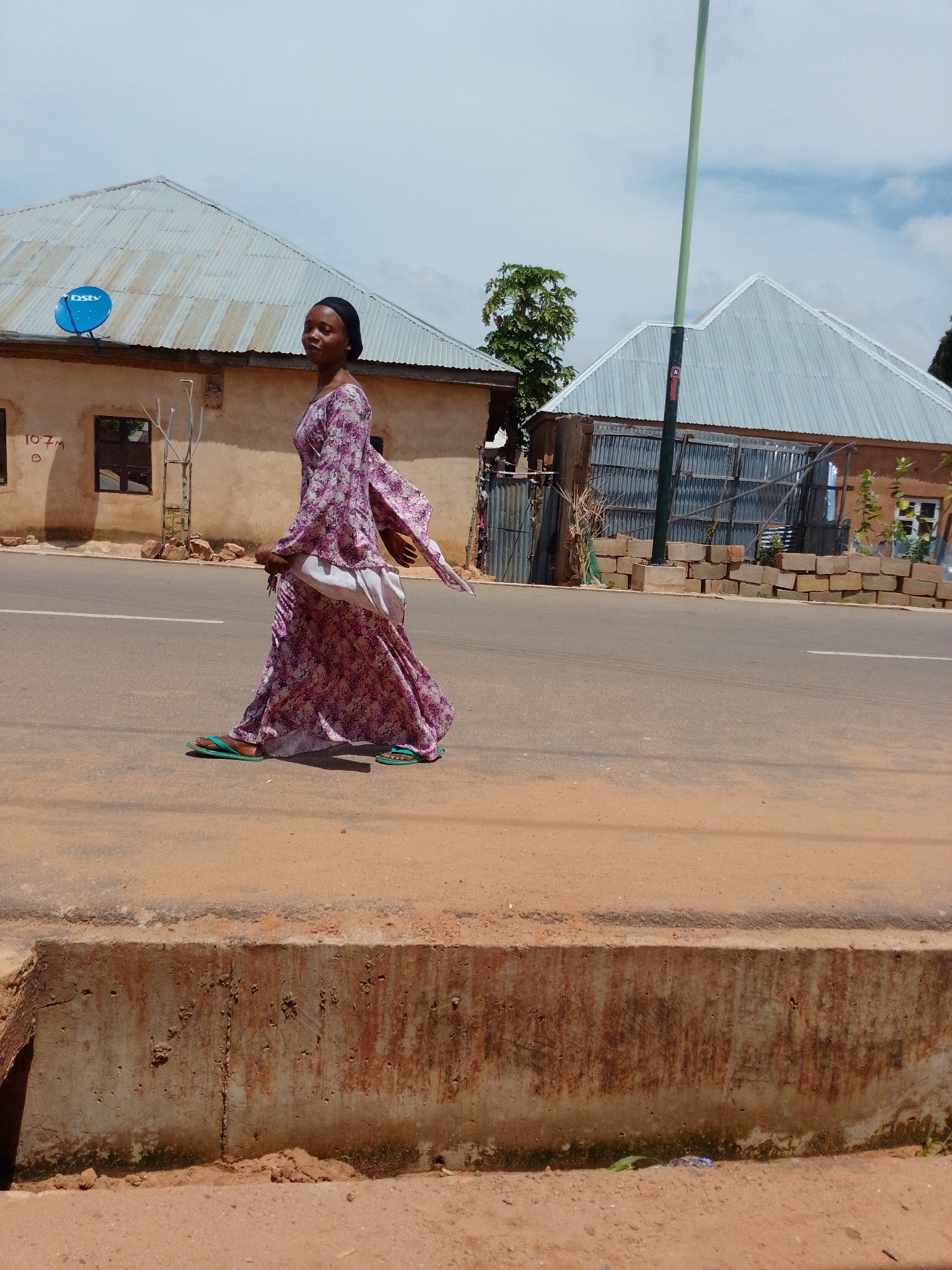 A woman wearing a floral print dress and a headscarf walks on the side of a paved road in front of a building with a corrugated metal roof. She is walking to the left of the frame. The building behind her is made of light-colored stucco and has two dark windows. A blue satellite dish is mounted on the roof of the building. A green utility pole stands in the background. The ground in the foreground is dusty and there is a concrete barrier along the edge of the road. The sky is partly cloudy.
