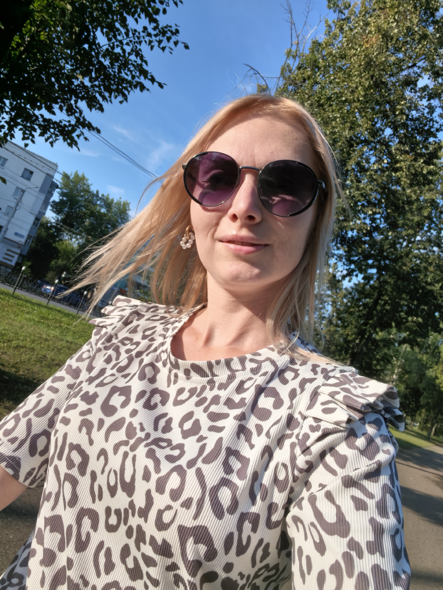 A blonde woman wearing sunglasses and a leopard print top is captured in a selfie outdoors. She has a pearl earring in her visible ear. The background shows a street with trees and a building, suggesting an urban park or residential area. The lighting indicates it is daytime with a clear blue sky. The location is identified as Sterlitamak, Russia.