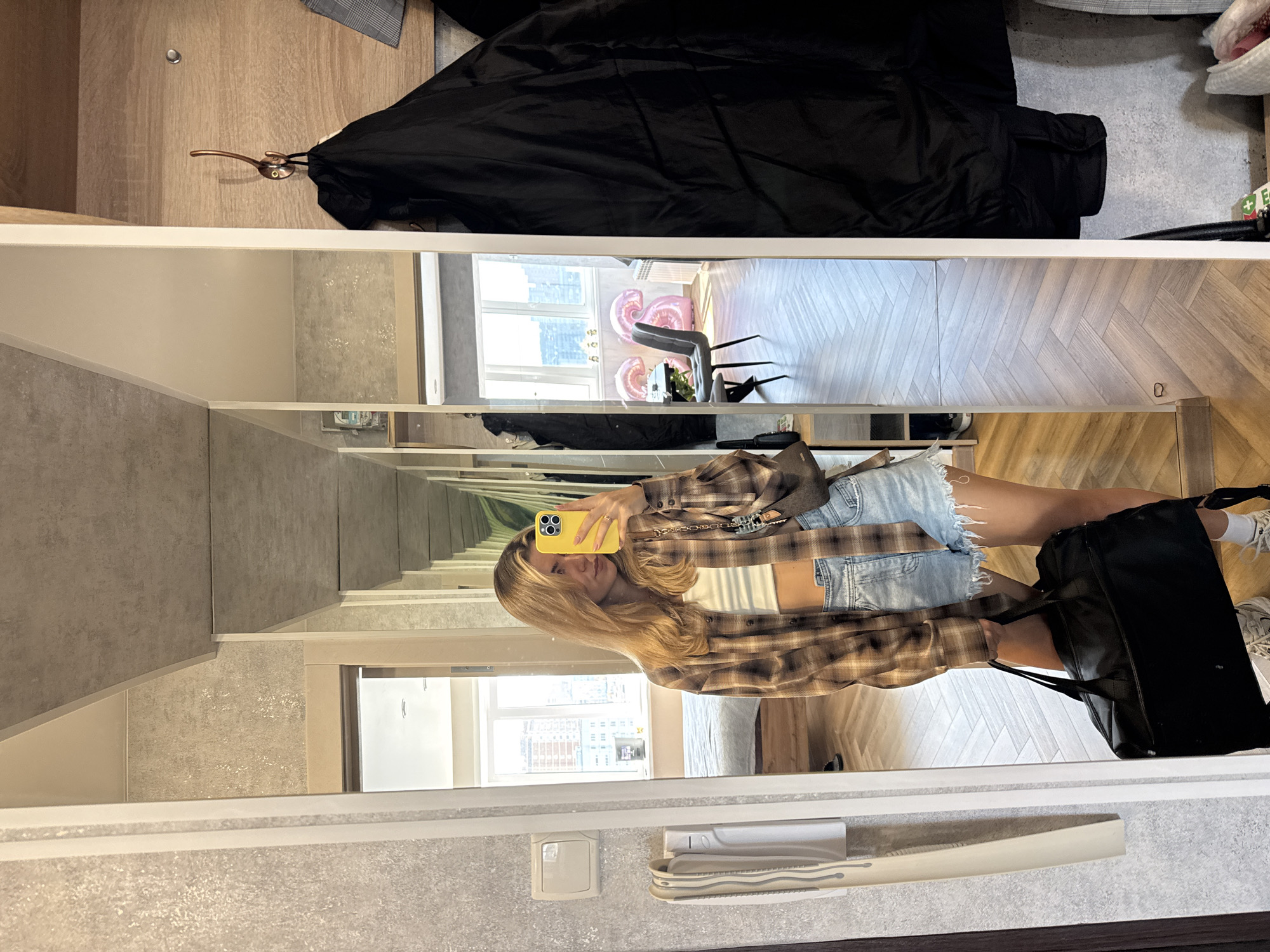 A mirror reflection shows a young woman taking a selfie with a yellow smartphone. She is wearing a plaid shirt, white top, and denim shorts. She is holding a large black bag. The reflection reveals a room with hardwood floors and a window with balloons and furniture. The location is city of Unknown, Unknown.