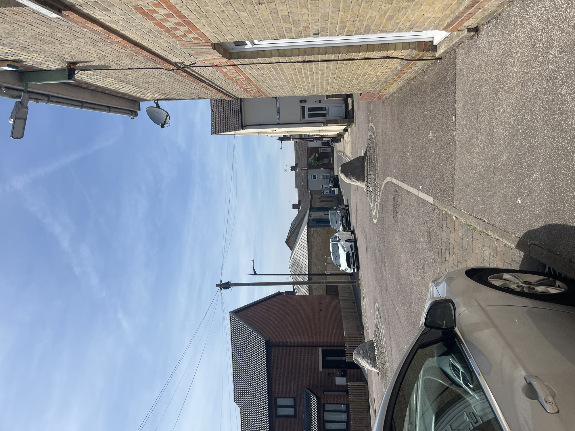 This image depicts a residential street in East Suffolk, United Kingdom. The view is taken from street level, looking down the road. The street is narrow, flanked by brick buildings on both sides. One side of the street has a sign reading "Cambridge Street". A silver car is partially visible on the right side of the frame, parked along the street. Further down the street, other parked cars are visible. The road surface is asphalt, marked with white lines. Periodically, there are raised stone planters at the road side with what appears to be wireframe surrounding. Overhead, power lines run along the street, supported by utility poles. The sky is blue with scattered clouds. A surveillance camera and a satellite dish are mounted on the side of one of the buildings.