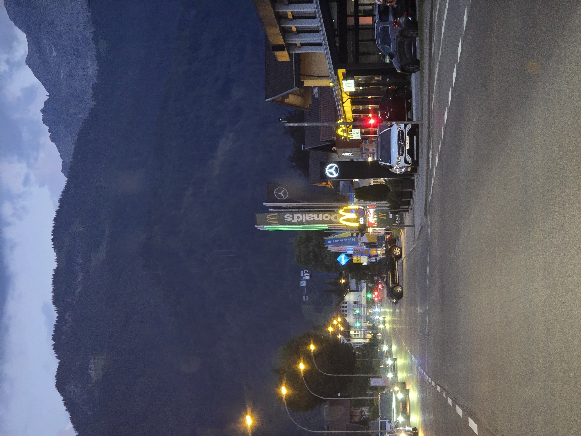 The image depicts a street scene at dusk or night in Garmisch-Partenkirchen, Germany. The perspective is from a low angle, looking down a road lined with buildings and illuminated by streetlights and signs. A prominent McDonald's sign with neon green lettering is visible, along with a Mercedes-Benz logo. Cars are parked and driving on the road, their headlights and taillights creating streaks of light.
In the background, a dark, tree-covered mountain looms under a twilight sky with some clouds. The overall atmosphere is one of a commercial area transitioning into the night, with the artificial lights contrasting against the natural darkness of the landscape. The image appears to be taken from a slightly elevated position looking down the road. There are no people clearly visible in the image. The weather appears clear and calm. The time of day is likely evening, given the twilight sky and the illumination from the businesses and streetlights.