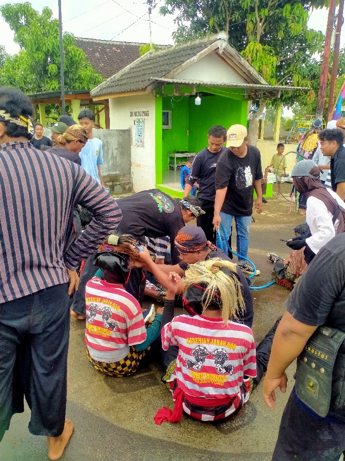The scene takes place in a residential area in Kecamatan Kedawung, Indonesia, at the coordinates -7.52 latitude and 111.029 longitude. A group of people are gathered around a man in a white and red striped shirt with the words "Kesehian Jarak, 0000000000" on it. The man is lying on the ground in front of a small building with green painted walls. Two other men in black shirts appear to be aiding him. There are multiple people around them, including one man in black and white striped shirt, several other men in black shirts, and a man in a white shirt with a red and black plaid scarf. There is a blue hose on the ground. The scene appears to be taking place in a street, with a small building and other residential structures visible in the background.