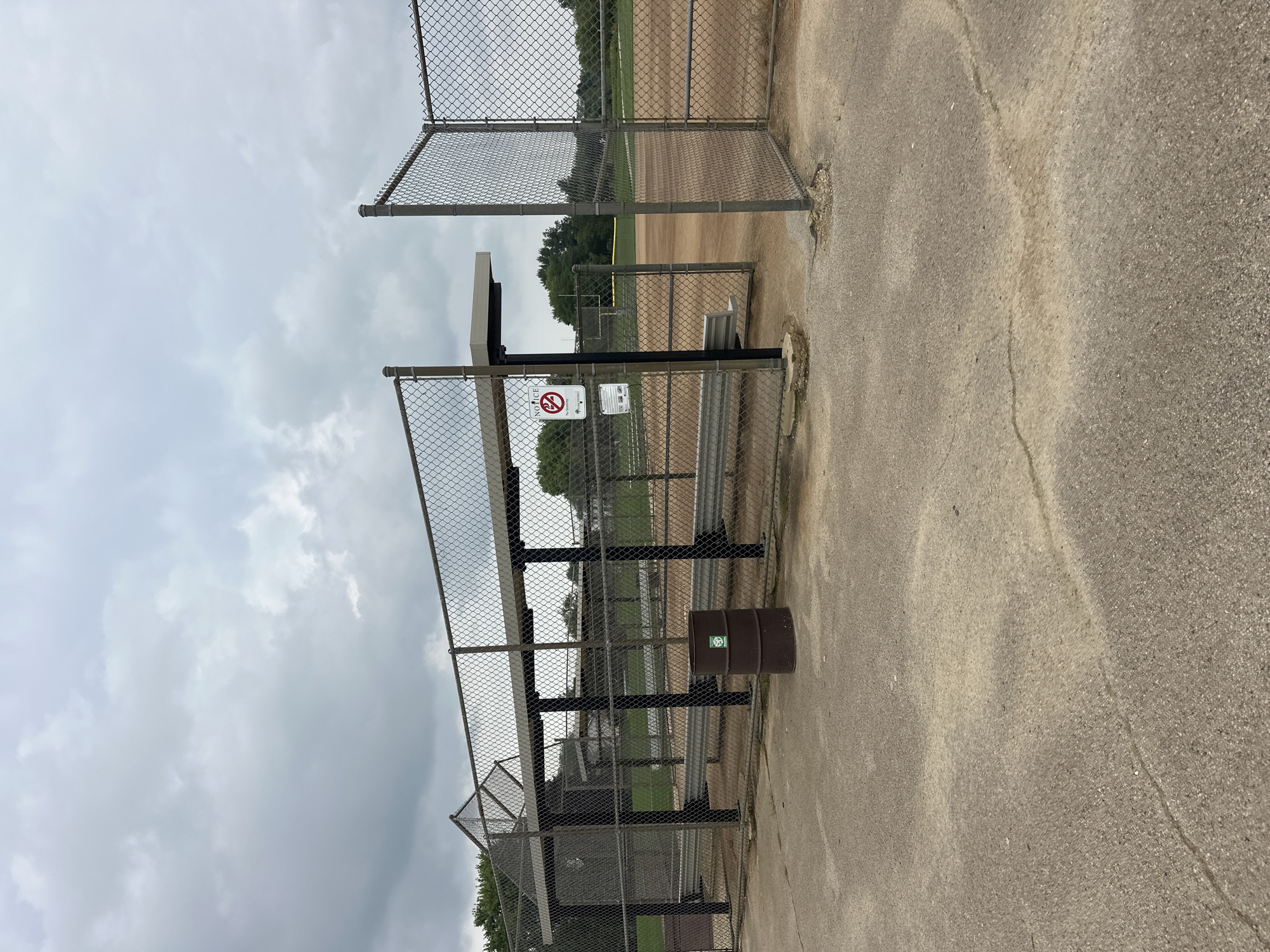 The image depicts a baseball field dugout with a chain-link fence enclosure in Schaumburg, Illinois, USA. The dugout features a metal roof supported by black poles, providing shade for a bench. To the left of the dugout, a brown metal barrel serves as a trash receptacle. A "Notice: No Smoking" sign is posted on the fence, along with a green and white circular emblem. The baseball field itself, composed of dirt and grass, is visible beyond the fence. The sky is overcast with gray clouds. The ground in front of the dugout is paved with concrete, showing some cracks and wear.