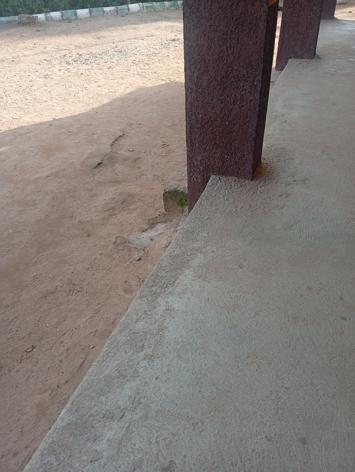 This image captures an outdoor scene in Jalingo, Nigeria, likely during the daytime with visible shadows suggesting sunlight. The primary subjects are architectural elements and the surrounding environment. A dark reddish-brown, textured pillar stands prominently in the foreground, suggesting it's part of a covered walkway or building. To the right of the pillar, a light gray concrete surface slopes downwards, forming a pathway or veranda.
Behind the pillar and to the left, the ground is composed of sandy, dusty earth, with some scattered small stones and debris. A faint crack or line is visible in the dirt. In the background, a low border wall with alternating green and white sections runs horizontally, separating the dusty area from a band of green foliage, likely trees or bushes. The overall impression is one of a simple, functional outdoor space, possibly a public area, school, or residential compound. No people or specific activities are depicted; the focus is on the static elements of the setting. The textures of the concrete, pillar, and dirt are clearly visible. No text is discernible in the image.