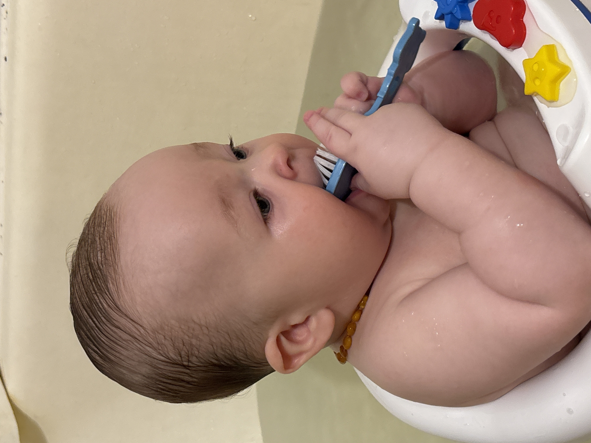 A baby is in a bathtub, sitting in a baby bath seat. The baby is fair-skinned with wet, dark hair. The baby has a amber-colored necklace. They are holding a blue toothbrush in their mouth. The baby bath seat has a yellow star, a red heart and a blue flower. The background is a beige-colored wall. The image was taken in Марковский, Russia.