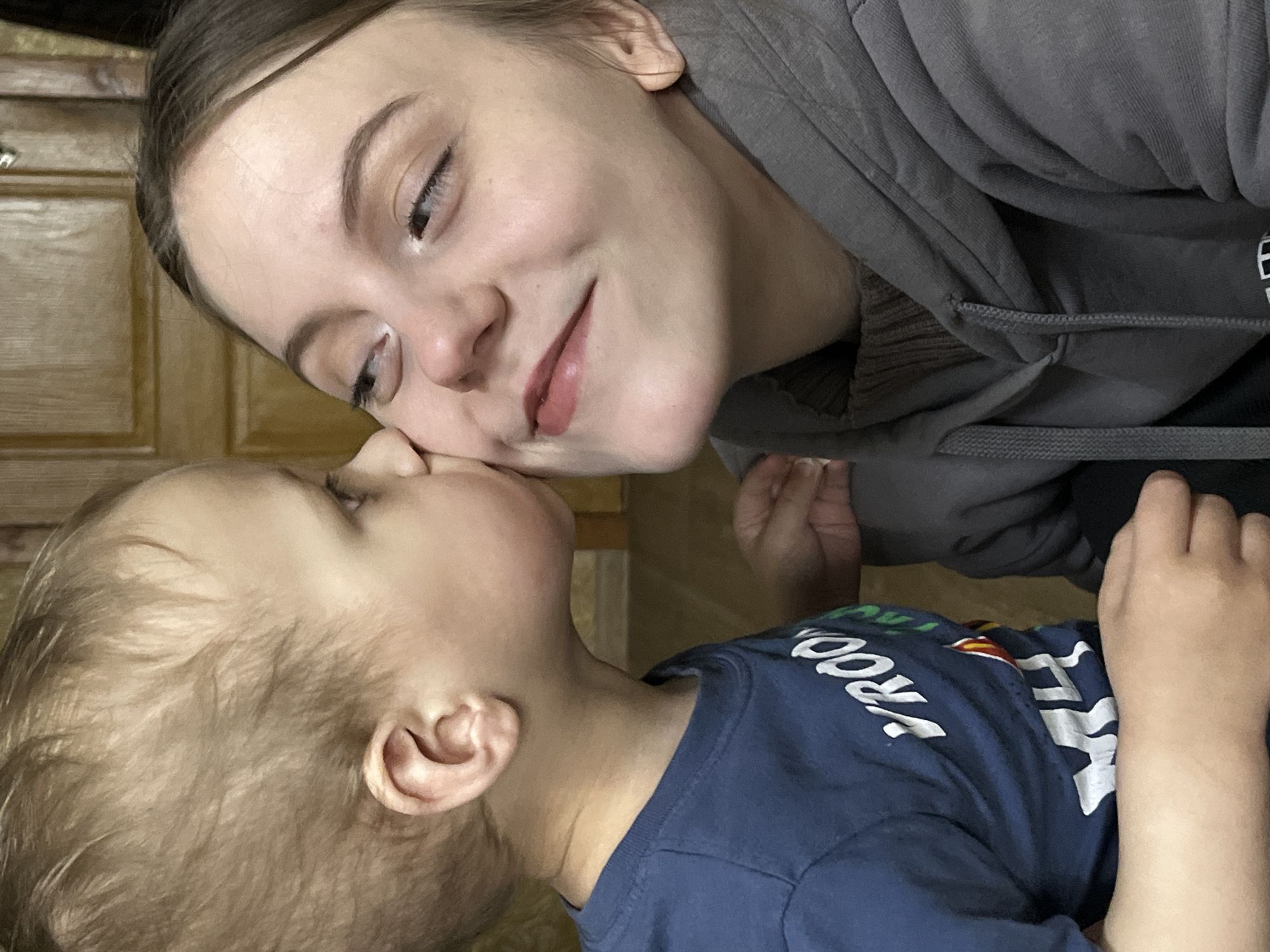 This indoor photograph captures a tender, affectionate moment between an adult female and a young child in what appears to be a home setting, possibly in Фоки, Russia. The image is taken from a slightly tilted perspective, focusing intimately on the faces of the subjects.
The adult, positioned in the upper portion of the frame, has light brown hair pulled back and is wearing a grey hooded sweatshirt with ribbed cuffs. She is leaning down, her face turned towards the child with a gentle, soft smile, and her eyes, accented with subtle eyeliner, convey warmth and affection. Her lips have a natural, light pink tint.
The young child, situated in the lower part of the frame, has light blonde or light brown hair and is wearing a navy blue t-shirt with white lettering. Visible text on the shirt includes "TROO" and "A L", suggesting part of a word like "MAROON". The child is looking upwards towards the adult, their faces very close, almost touching, which emphasizes their intimate bond. The overall emotion conveyed is one of love, happiness, and a strong familial connection.
The background features light brown wooden cabinetry or furniture, indicating a domestic environment. The lighting suggests it is daytime, though no direct light source is visible.