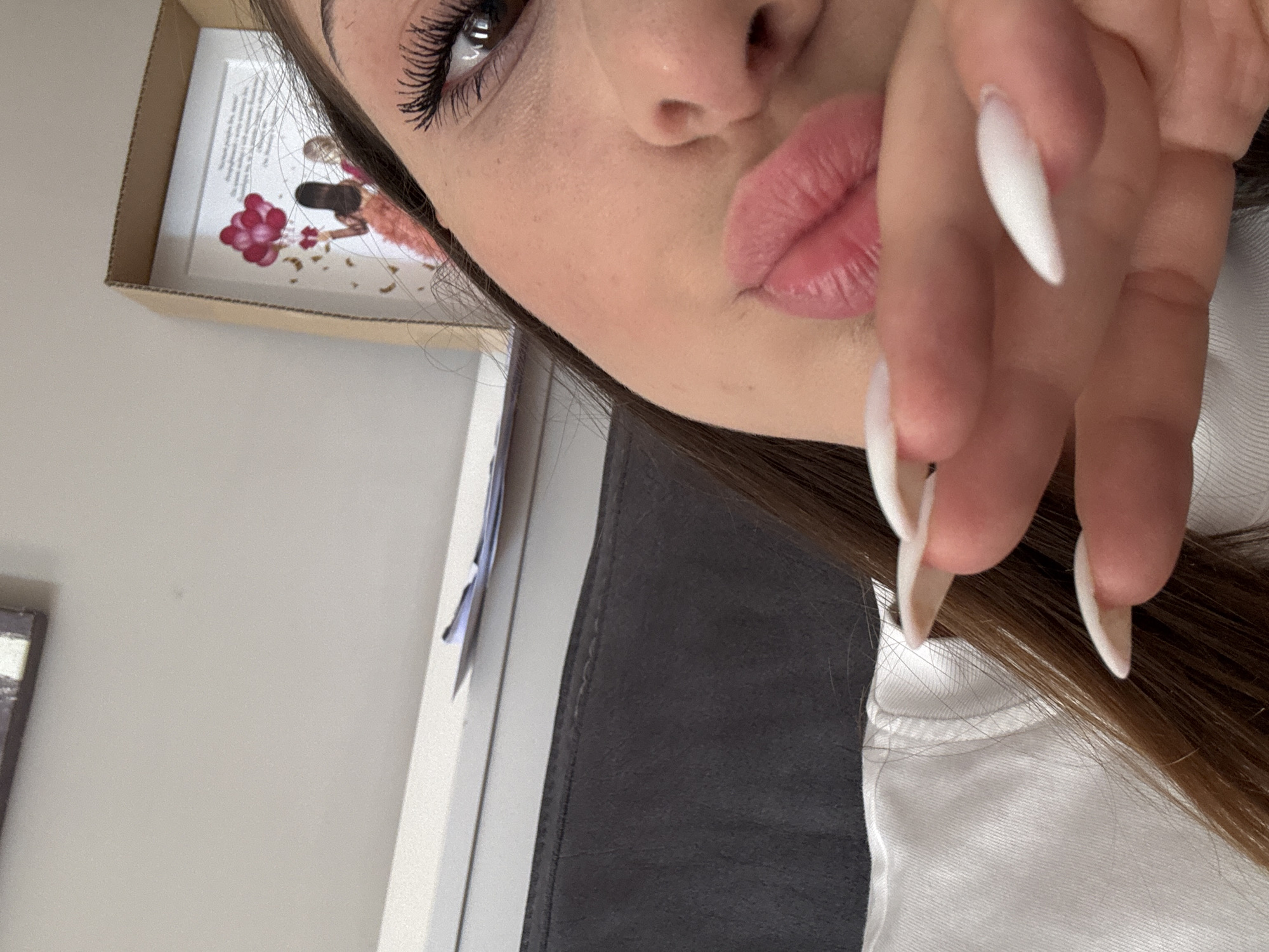 A close-up, shallow depth of field image shows the right side of a person's face, including their eye and lips, with their hand positioned in front of their mouth. The person has dark hair, dark eyelashes, and is wearing makeup. Their lips are slightly parted and appear to be a pinkish-red color. The hand has long, white, almond-shaped fingernails. In the background, a framed picture with two women and balloons is visible, along with a white shelf and a dark gray surface. The environment appears to be indoors, possibly a room in a home.