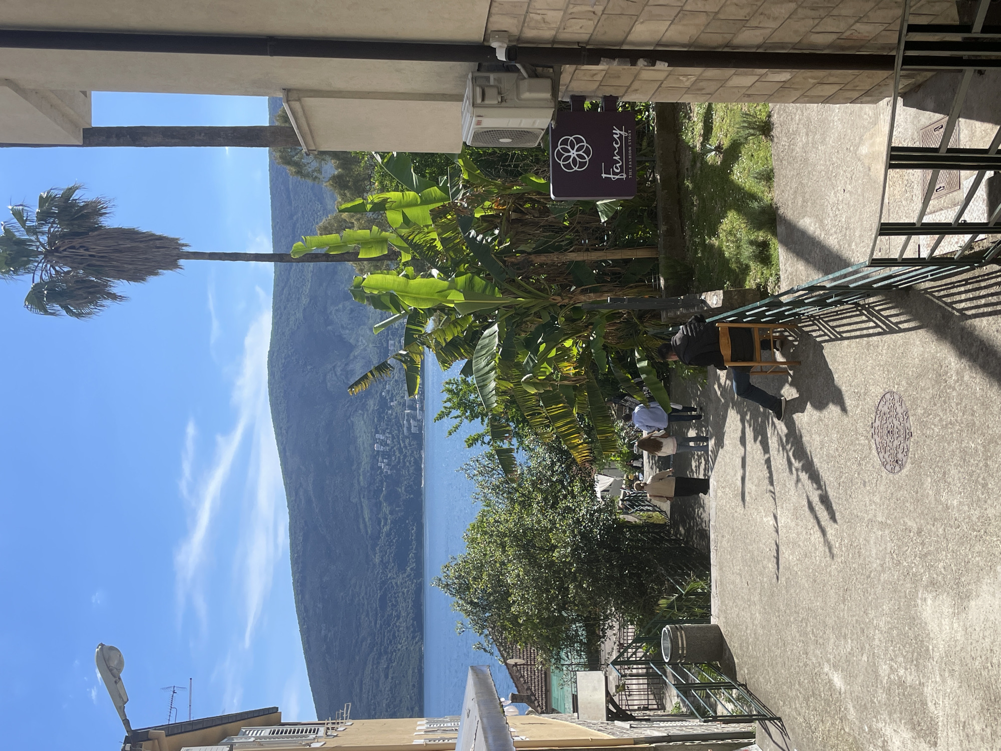 A vertical shot shows a scenic view from a building in Herceg Novi, Montenegro. The landscape features a large body of water with a backdrop of mountains on a sunny day. The sky is mostly clear blue with some wispy clouds.
In the foreground, a view from within a building is framed by parts of the structure at the top and bottom of the image. A tall palm tree is visible to the left, its leaves extending into the sky. The camera position looks to be from a balcony, as a metal railing runs along the right side of the frame.
In the middle ground, a group of people appears to be standing and sitting in an outdoor area. Lush green vegetation, including banana trees, is prominent. A sign for "Fancy - The Fashion Story" is visible among the trees.