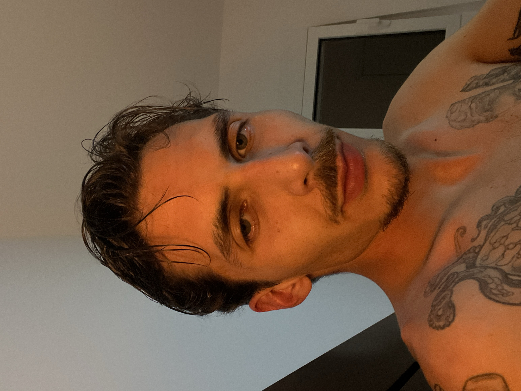A close-up shot of a man's face and upper torso. He has brown hair, hazel eyes, and a mustache and goatee. His hair appears wet. He has tattoos on his right shoulder and chest. The background consists of a white wall and a window with a dark frame. The location of the image is not inferable from the given information.