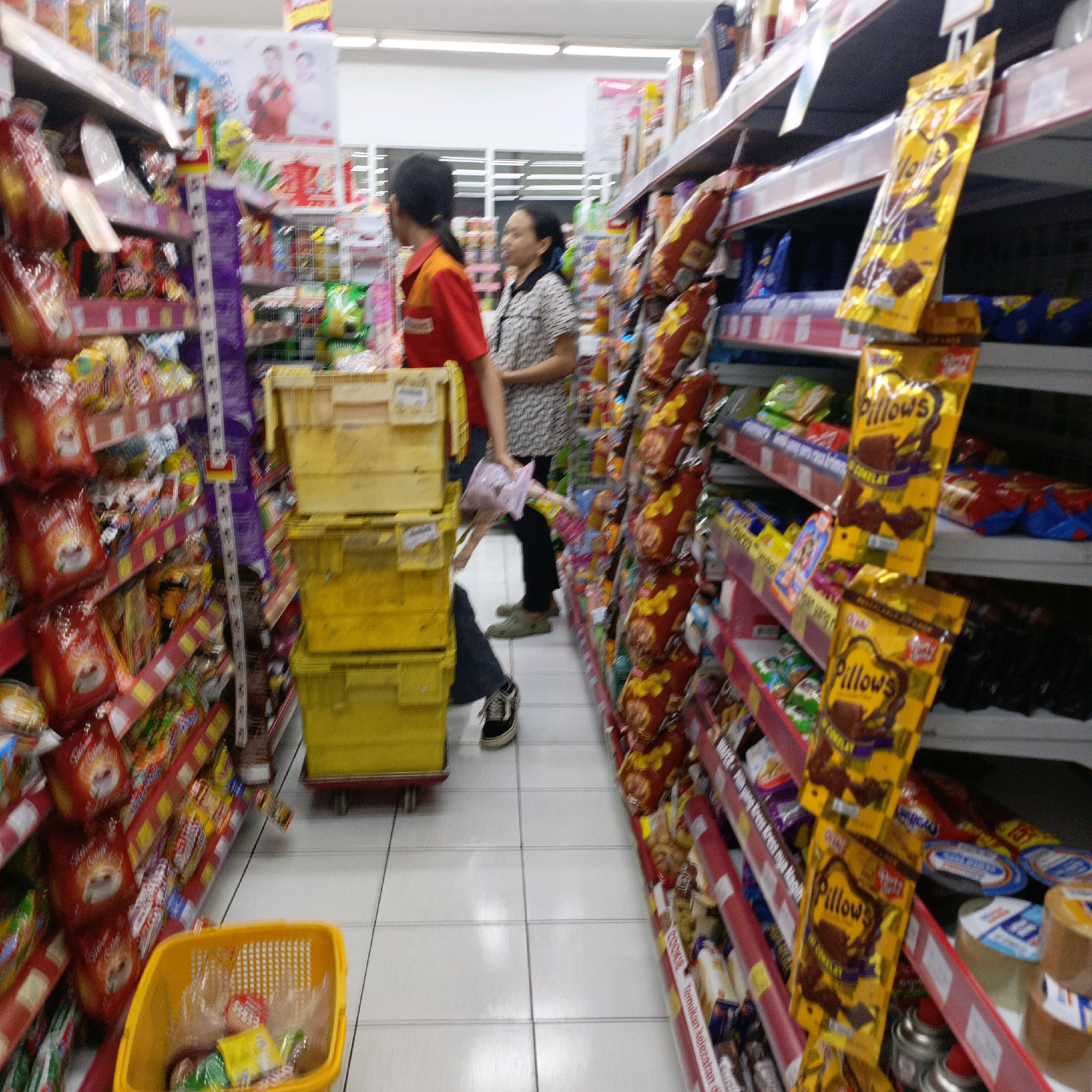 The image depicts the interior of a retail store, likely a supermarket or minimarket, in Salatiga, Indonesia. The scene focuses on aisles stocked with various packaged goods, primarily snacks and food items. In the foreground, a bright yellow shopping basket is visible, containing some plastic-wrapped items. To the left, shelves are filled with brightly colored packages, many with red and gold hues.
In the middle ground, two individuals are present. A person wearing a red and orange uniform, possibly an employee, is standing near a stack of yellow plastic crates. Another person, a woman in a patterned shirt, is looking towards the right side of the frame. Behind them, more shelves are laden with products, including prominent yellow packaging with the word "Pillows" on them.
The floor is tiled with a light-colored material. The lighting appears to be artificial, typical of an indoor retail environment. The overall impression is a busy shopping aisle with goods displayed for sale. The presence of specific product packaging and the context of a retail environment strongly suggest this is a commercial establishment in Salatiga.