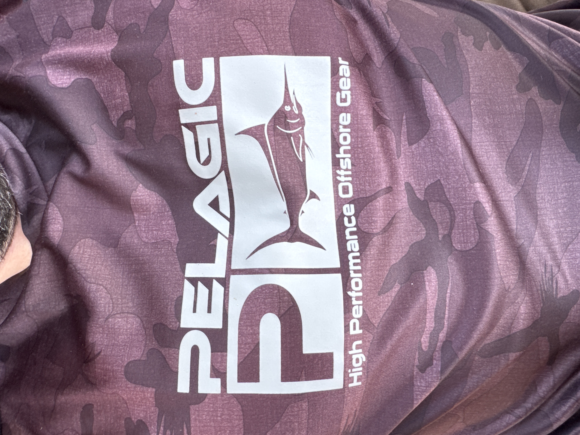A close-up, medium shot shows the front of a dark, camouflage-patterned shirt. The shirt has a graphic design on it. The top of the graphic features the word "PELAGIC" in large, white, sans-serif capital letters. Below "PELAGIC" is a white rectangle containing a stylized illustration of a marlin. To the left of the marlin is a large, white letter "P". Below the rectangle, also in white sans-serif capital letters, are the words "High Performance Offshore Gear." The shirt appears to be made of a synthetic, moisture-wicking fabric. The background is out of focus and shows folds and wrinkles in the fabric, suggesting it is being worn.