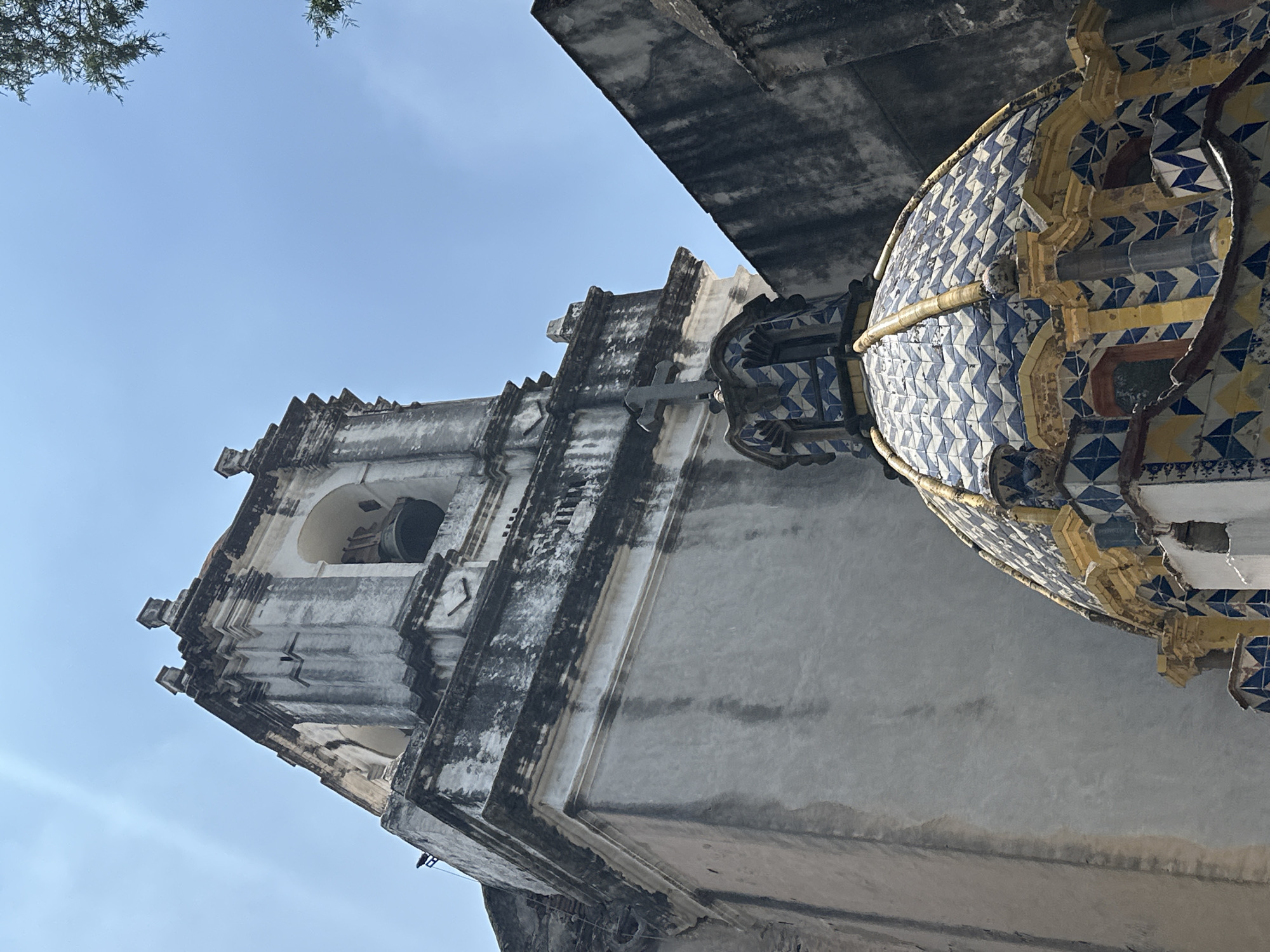 The image shows a low-angle view of architectural structures in Mexico City, Mexico. A church bell tower is visible on the left, constructed of aged white and grey stone, and featuring a visible bell within the belfry. The structure is crowned with distinctive stepped architectural elements. Adjacent to the tower is a rounded dome or cupola covered in blue and white patterned tiles with yellow detailing. The background is filled with a light blue sky and a portion of a tree at the upper left edge. The overall impression suggests an aged, historic, and architecturally significant structure.